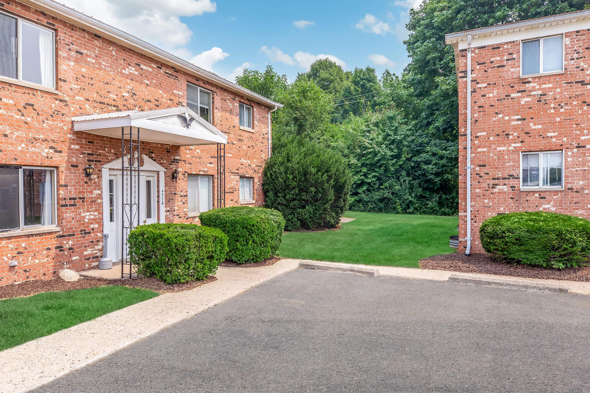 A view of a brick apartment complex featuring two buildings. The entrance has a canopy and is surrounded by neatly trimmed bushes. A grassy area is visible in the background, along with a clear blue sky and scattered clouds. The pathway leads to the entrance, emphasizing a well-maintained outdoor space.