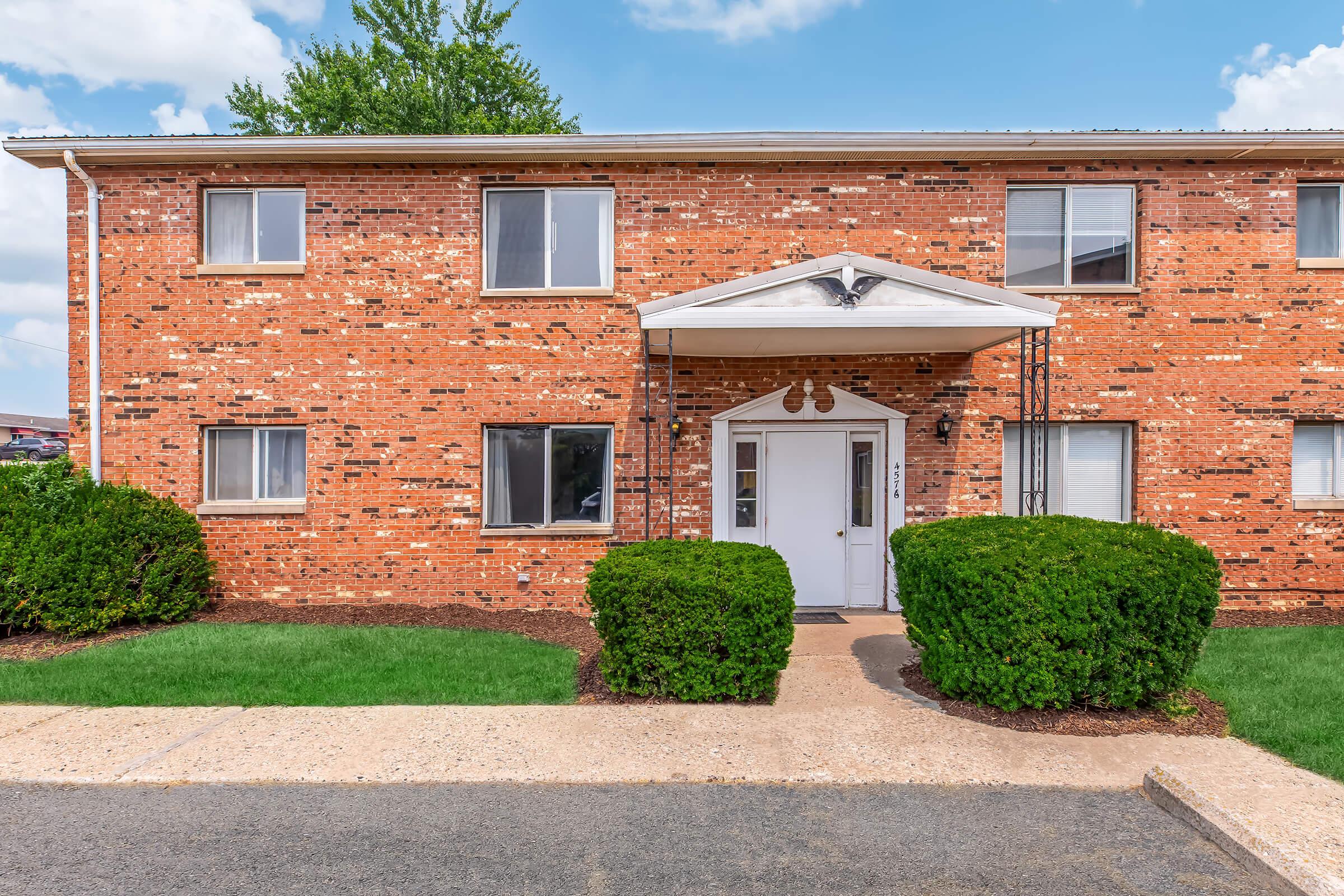 A two-story brick apartment building with a white entrance door and awning. The front yard features well-trimmed shrubs and a concrete pathway. The building has multiple windows, and the sky above is partly cloudy.