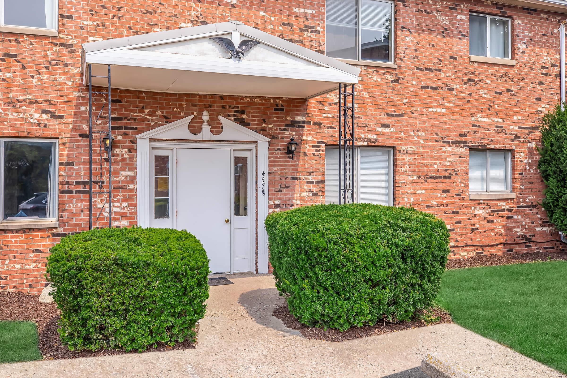 A brick exterior of an apartment building featuring a white front door under a small awning. Two neatly trimmed green shrubs flank the entrance, and a concrete pathway leads to the door. Windows are visible on either side of the entrance, with a well-maintained lawn in front.
