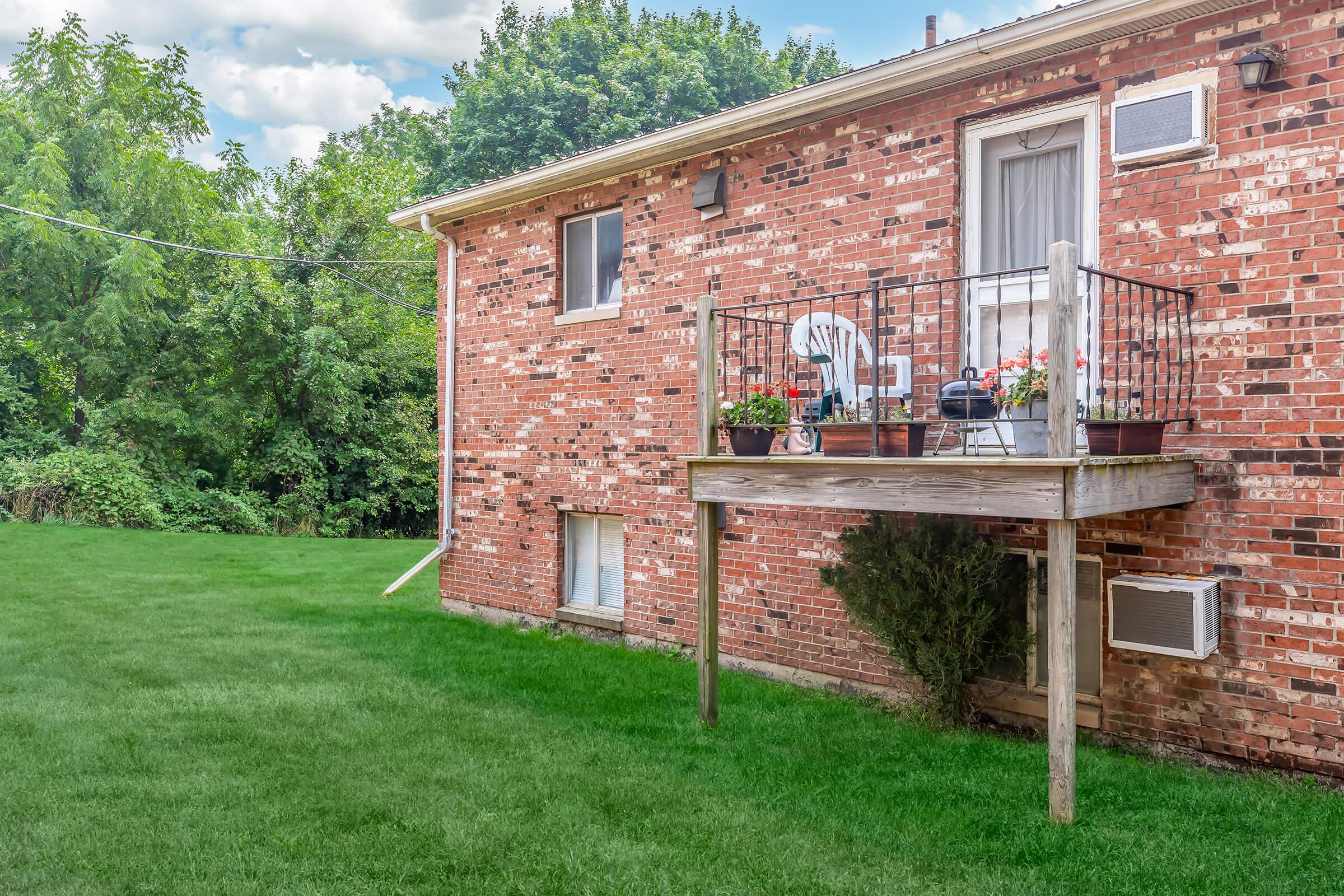 A brick building with a small wooden balcony on the lower level, featuring potted plants. There's a well-maintained green lawn in front, surrounded by trees and shrubs. The sky is partly cloudy, adding a pleasant ambiance to the scene.