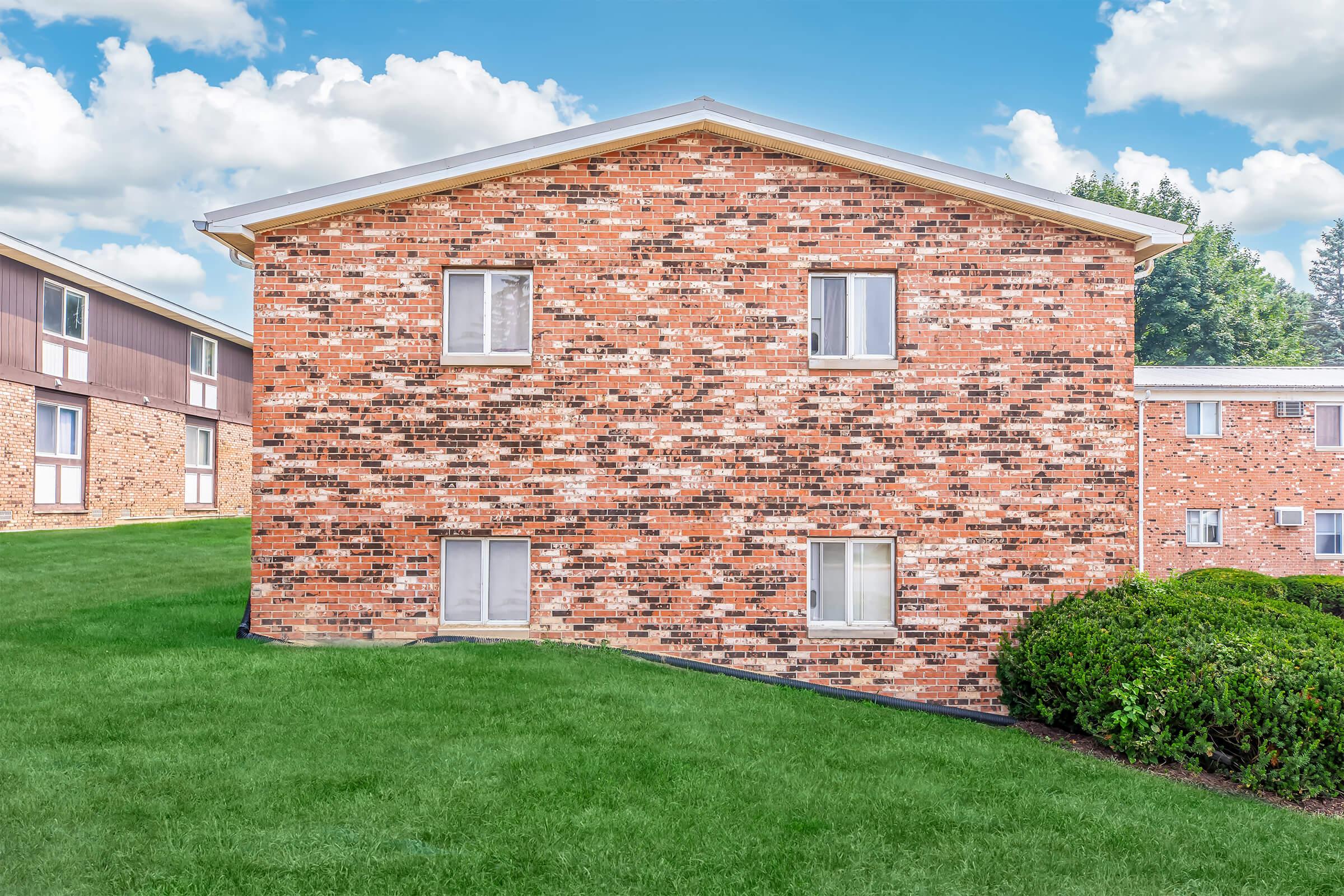 A brick apartment building with several windows sits on a well-maintained grassy area. The structure features a mix of dark and light-colored bricks, and the sky above is partly cloudy. Surrounding landscaping includes neatly trimmed bushes.
