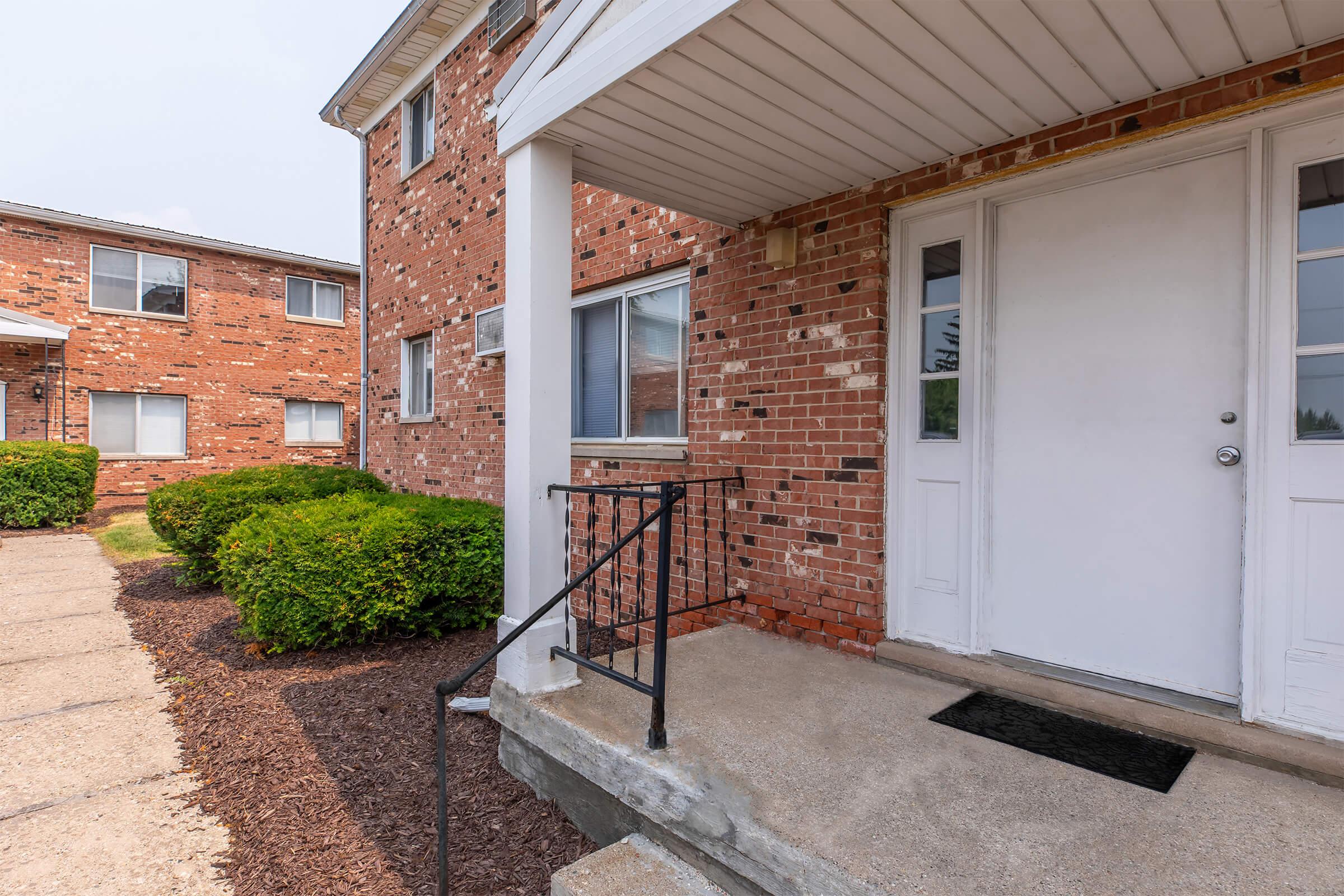 Entrance to an apartment building featuring a set of double doors with a small staircase. The building has a brick exterior, and there are neatly trimmed shrubs and a concrete walkway leading up to the entrance. Adjacent buildings are visible in the background.