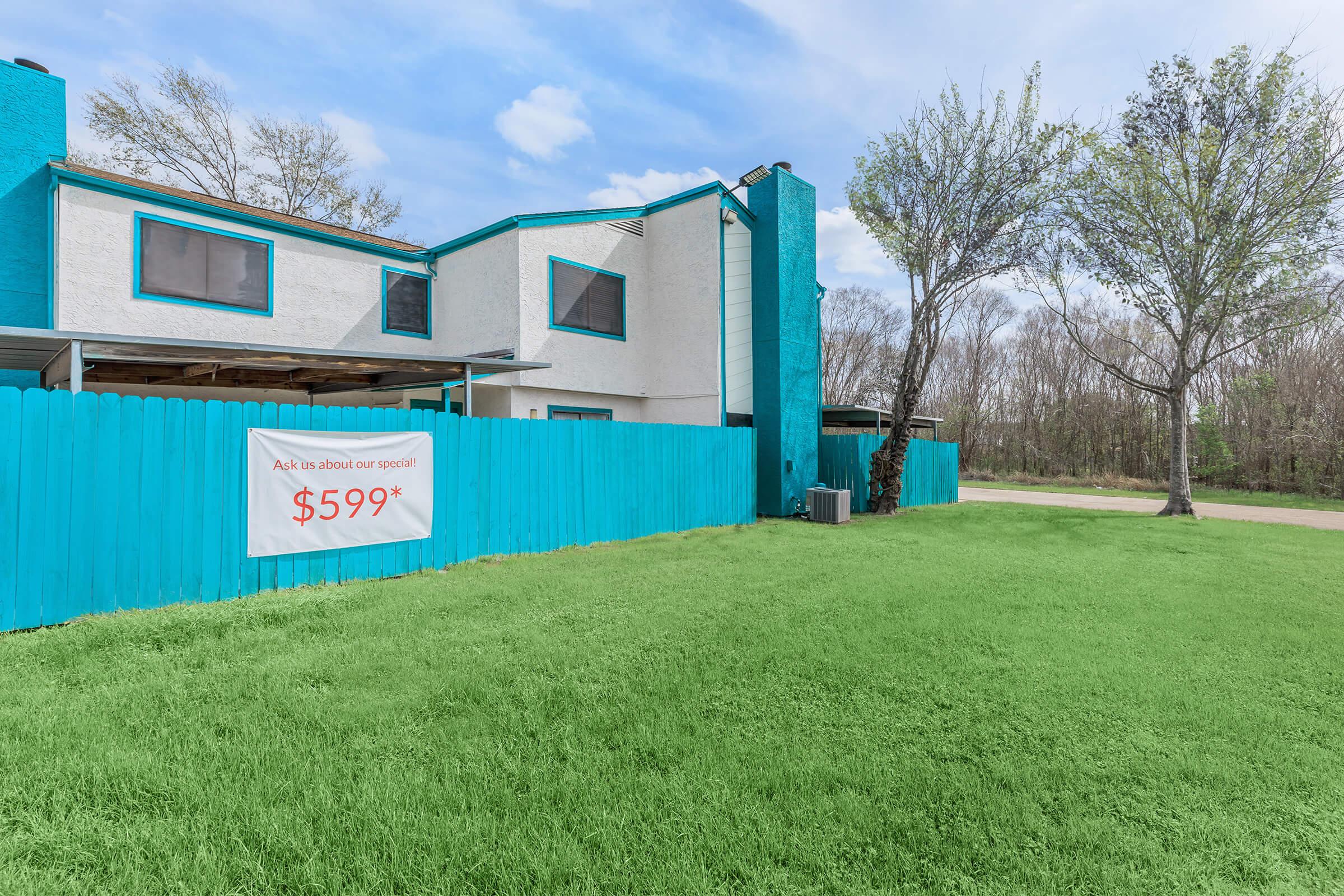 A two-story building with a bright turquoise and white exterior. There’s a large banner on a bright turquoise fence advertising a special price of $599. The foreground features lush green grass, and behind the building, trees with bare branches can be seen against a cloudy sky.
