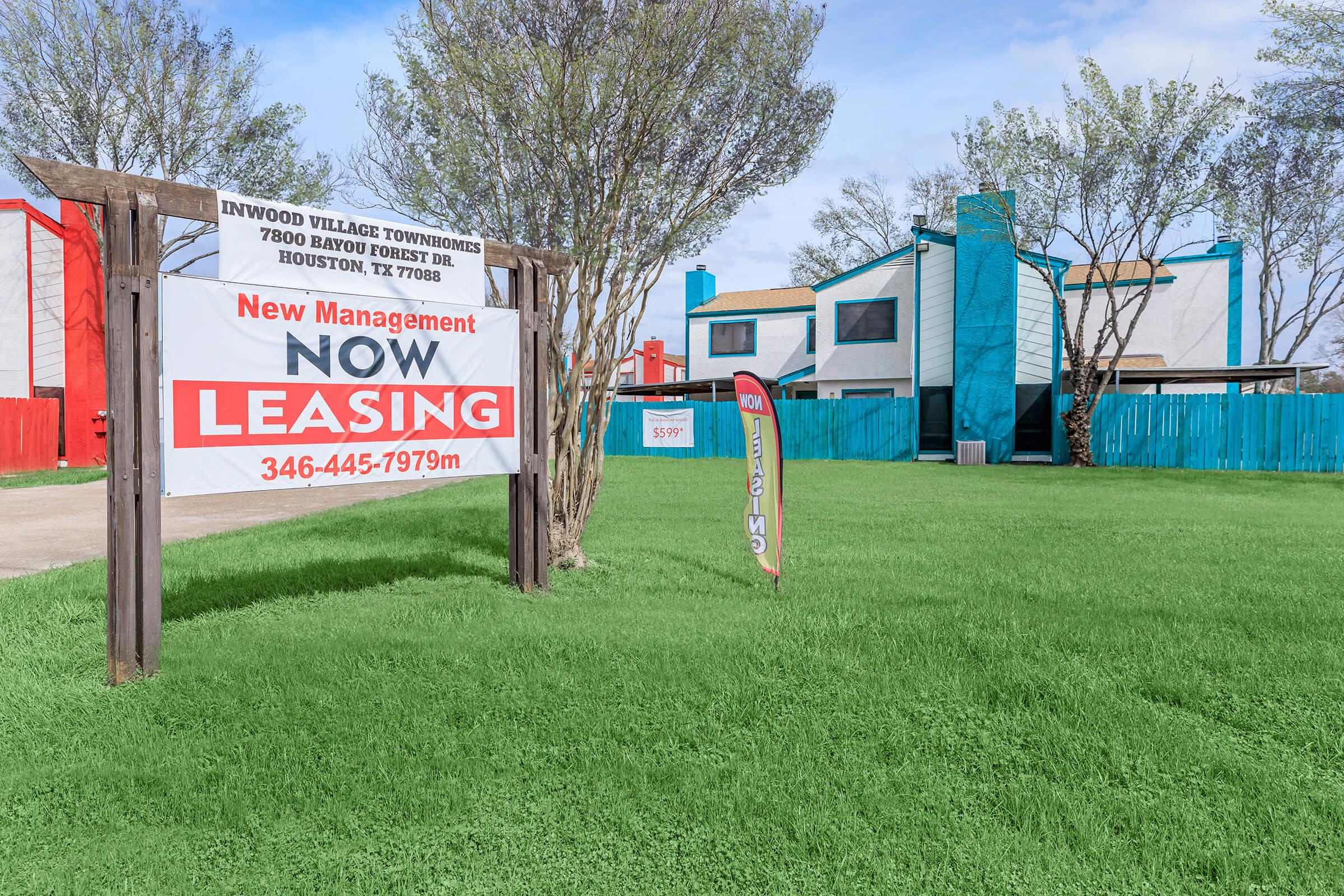 A colorful leasing sign in front of Inwood Village Townhomes in Houston, TX, featuring "New Management NOW LEASING" prominently displayed. The background shows a well-maintained grassy area with modern, multi-colored buildings. The image conveys an inviting property management message.