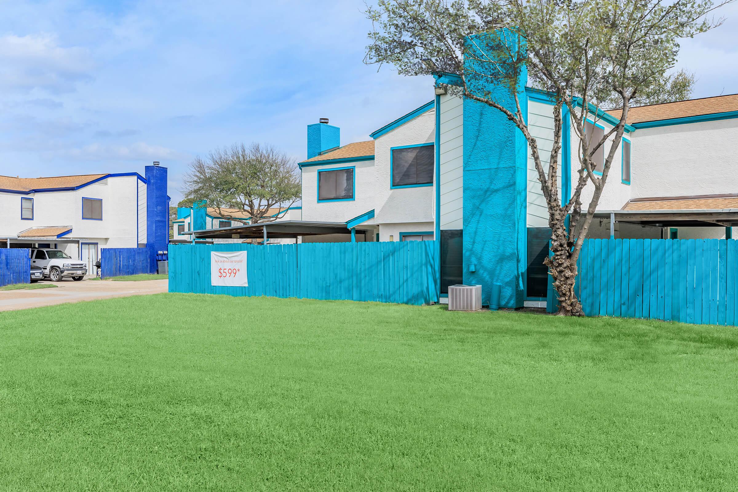 A colorful two-story building with a turquoise fence and accents, surrounded by a lush green lawn. A sign displaying "599" is visible on the fence. Nearby, there are additional blue structures and parked vehicles. The sky is partly cloudy.