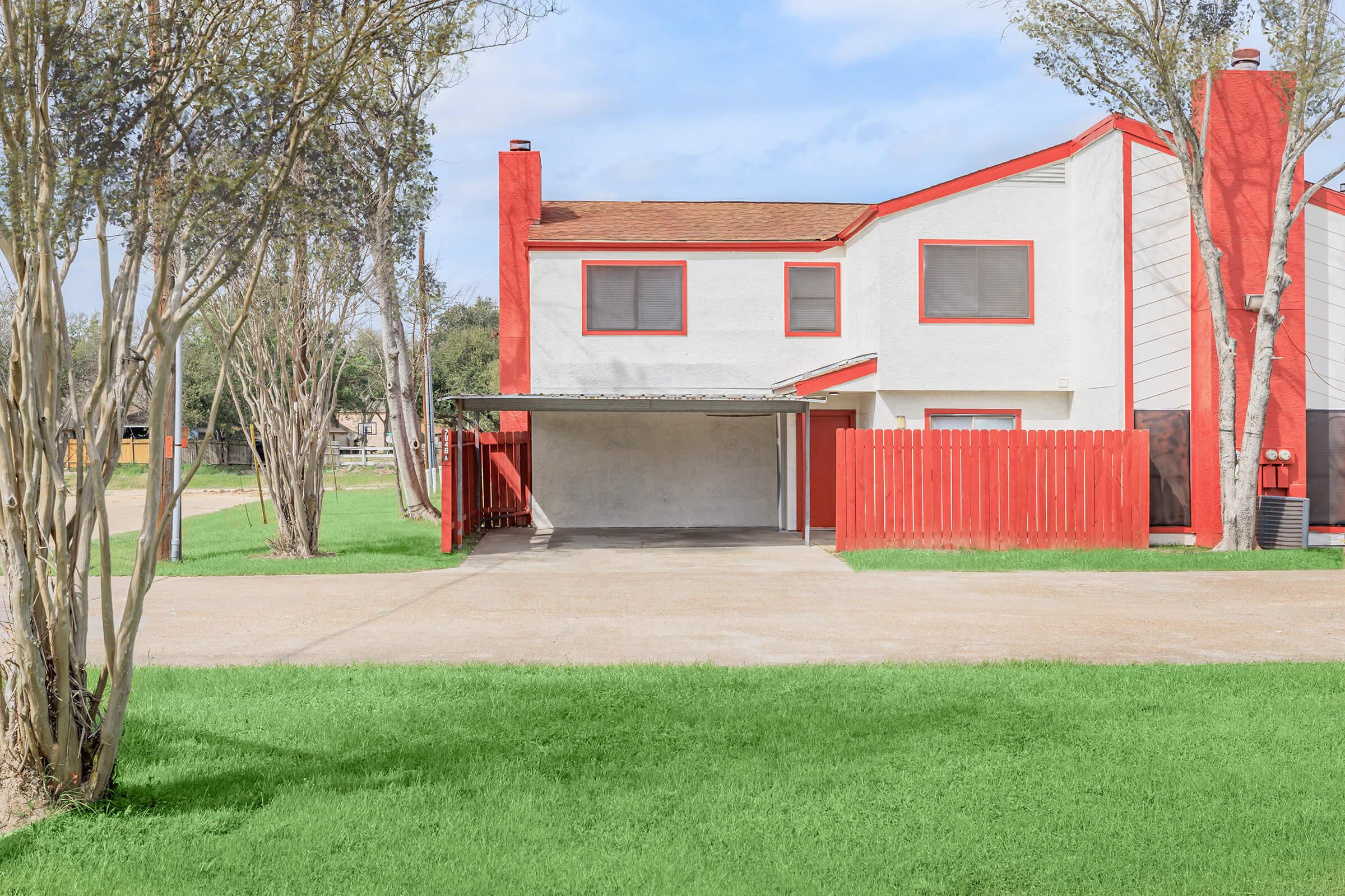 A two-story house with a red roof and accents, featuring a white exterior. There are two large windows on the upper floor, a covered carport, and a red wooden fence surrounding the property. Lush green grass surrounds the driveway, with a few trees in the foreground. The sky is partly cloudy.