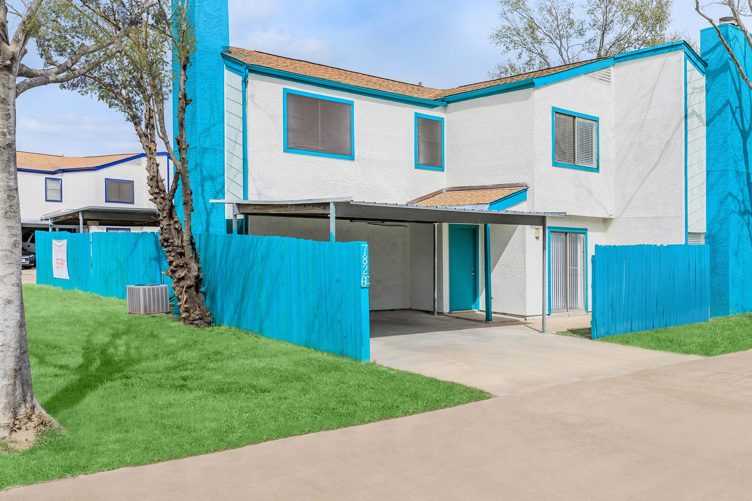Two-story residential building with white walls and bright blue trim. A carport area is visible, along with a grassy lawn leading up to the structure. The wooden fence around the carport is painted blue, complementing the building’s design. Trees are partially visible in the background under a clear sky.