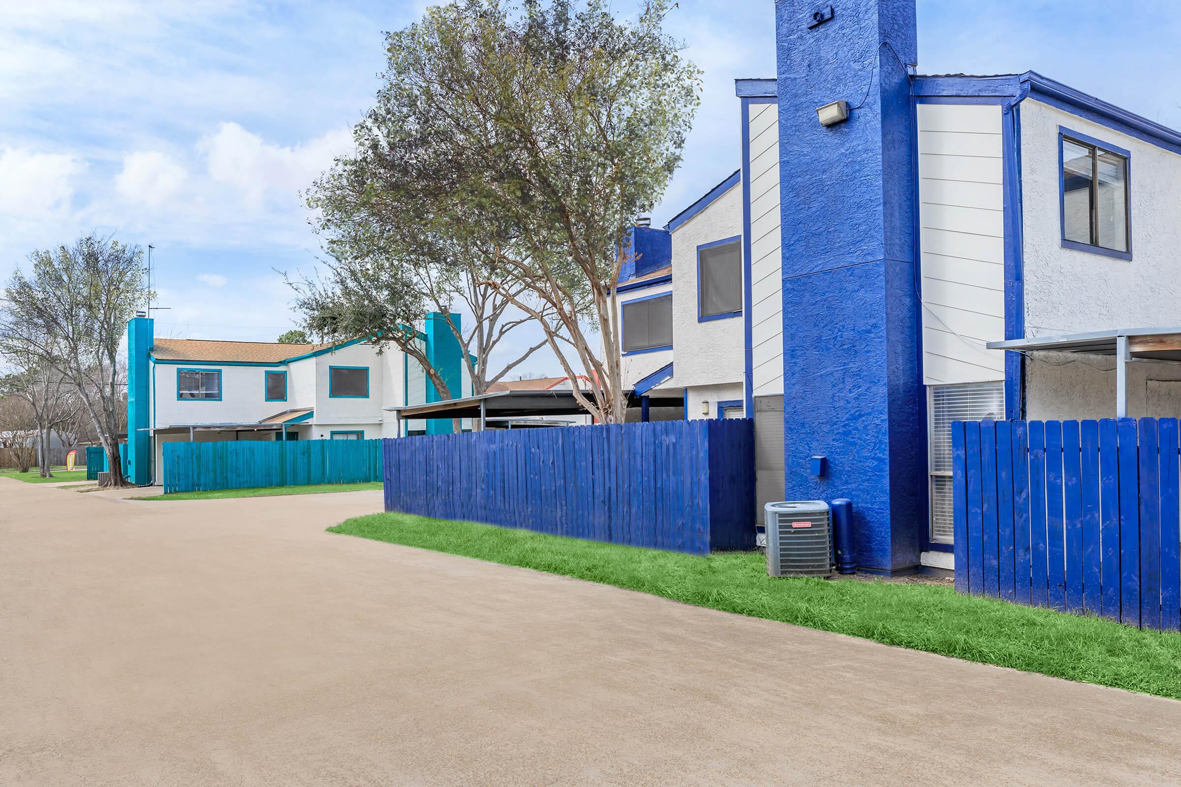 A view of a residential area featuring two buildings, one with blue accents and a white exterior. There's a blue wooden fence in the foreground, along with green grass and a tree. The pavement suggests a quiet street or pathway, creating a serene neighborhood atmosphere.
