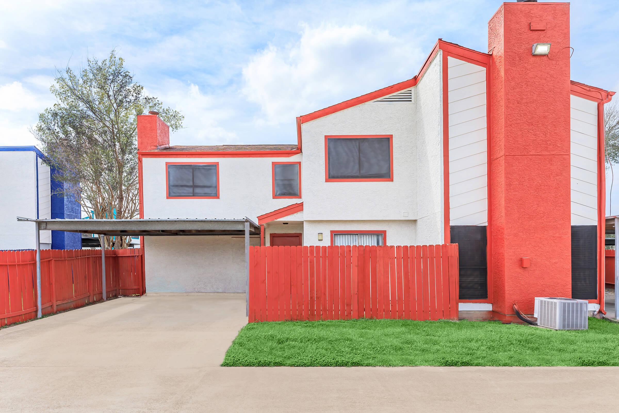 A modern two-story house featuring a mix of white and bright red exterior walls. It has several windows, a carport, and a small grassy area in front. The house is surrounded by a red wooden fence, and there are trees and a blue building visible in the background under a partly cloudy sky.