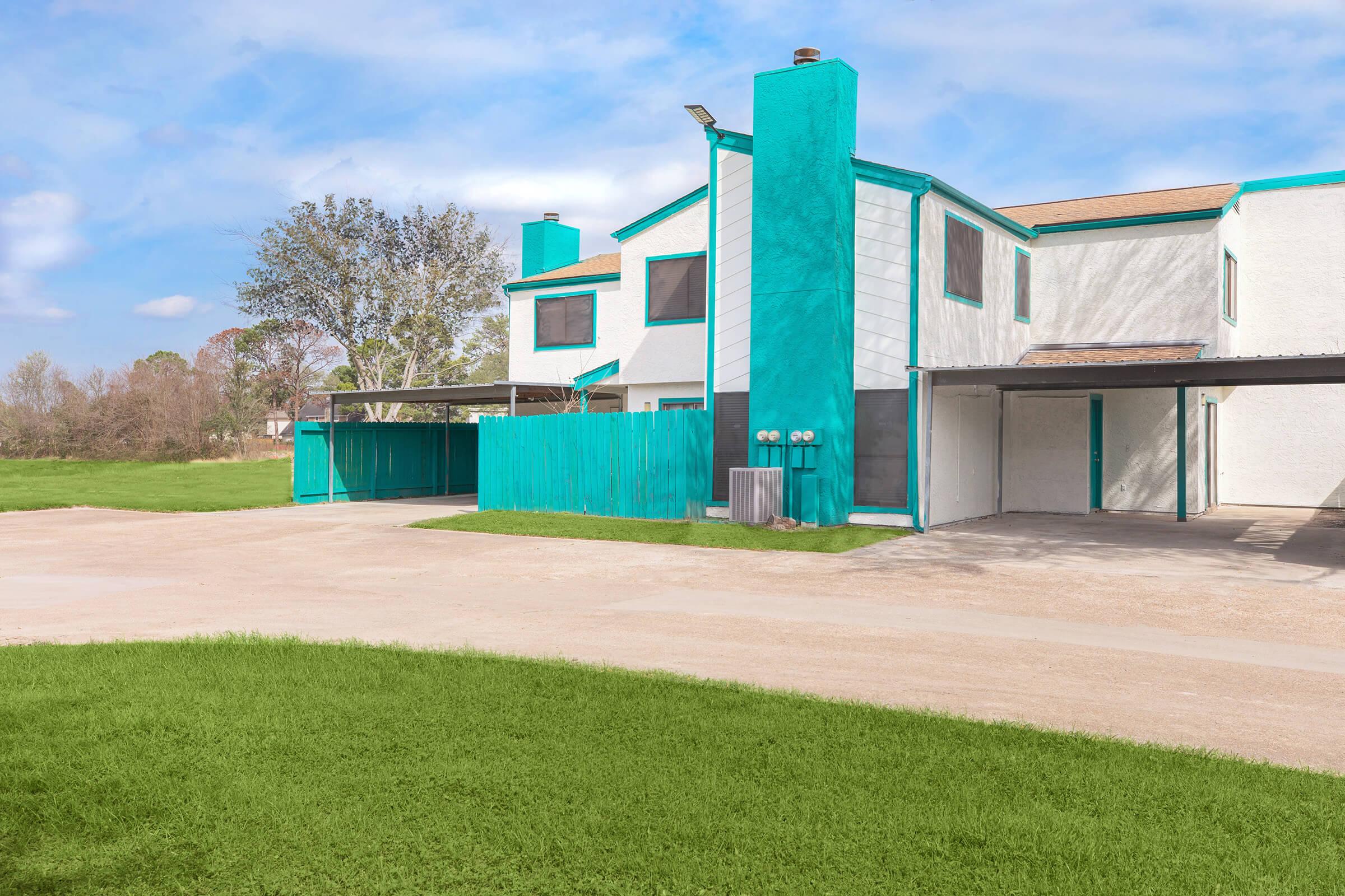 A modern two-story building with turquoise accents and white siding, featuring large windows and a covered parking area. The surrounding area has green grass and trees, with a clear blue sky in the background.
