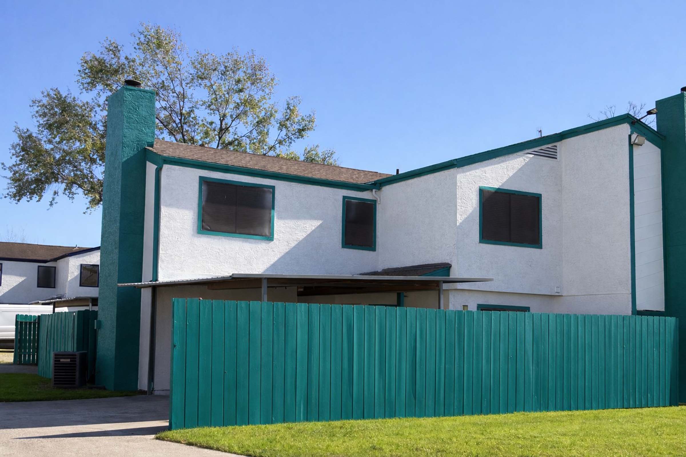 A two-story house with a white exterior, green trim, and a teal wooden fence. The building features a chimney and several windows, with a covered porch area. The surrounding area has a well-maintained lawn and clear blue skies.