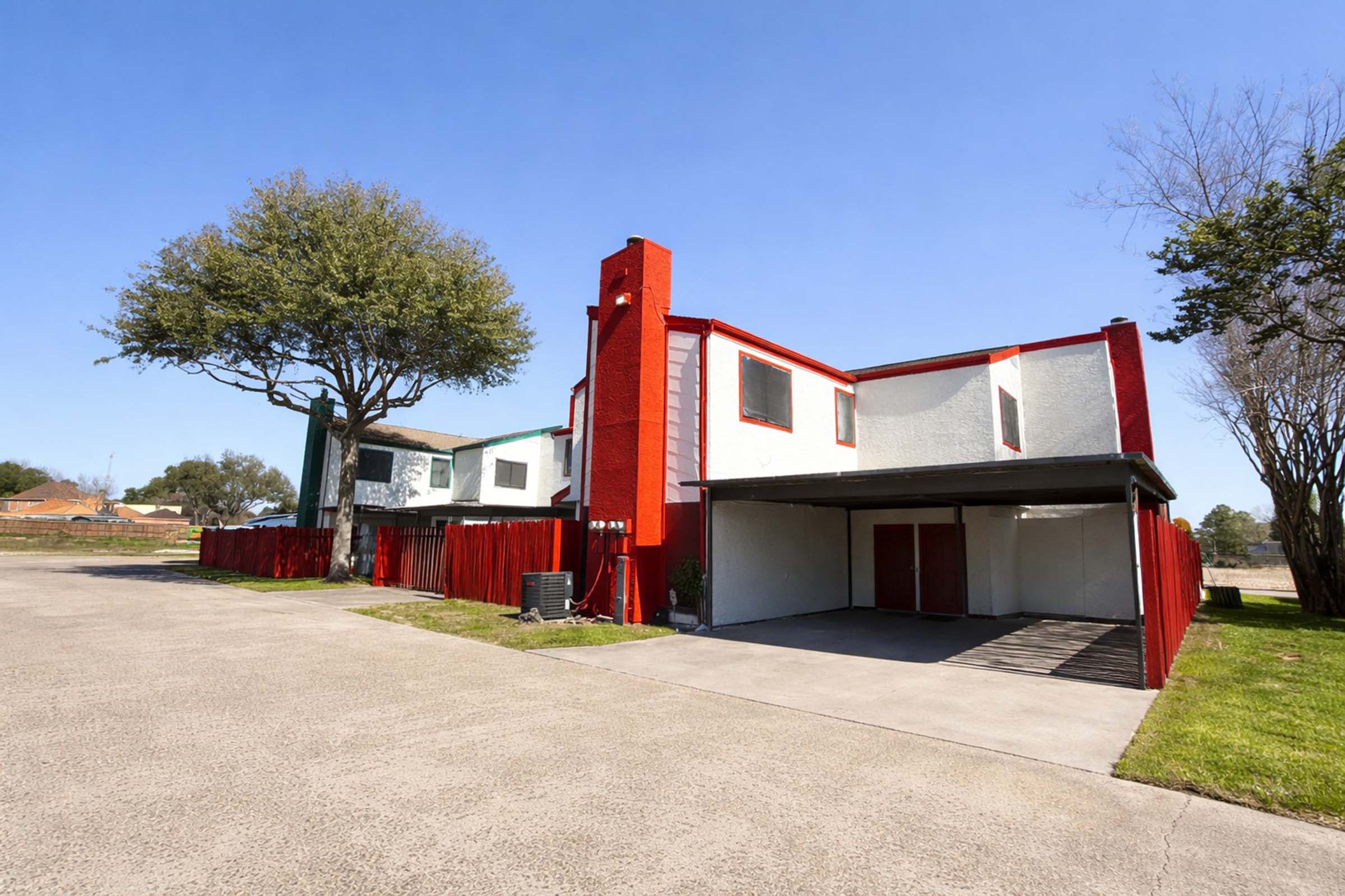 A modern two-story house painted in white and red, featuring a large tree nearby. The property has a concrete driveway and a covered parking area beneath the house. Clear blue skies create a bright atmosphere, and the surrounding area is open with grass and some trees.