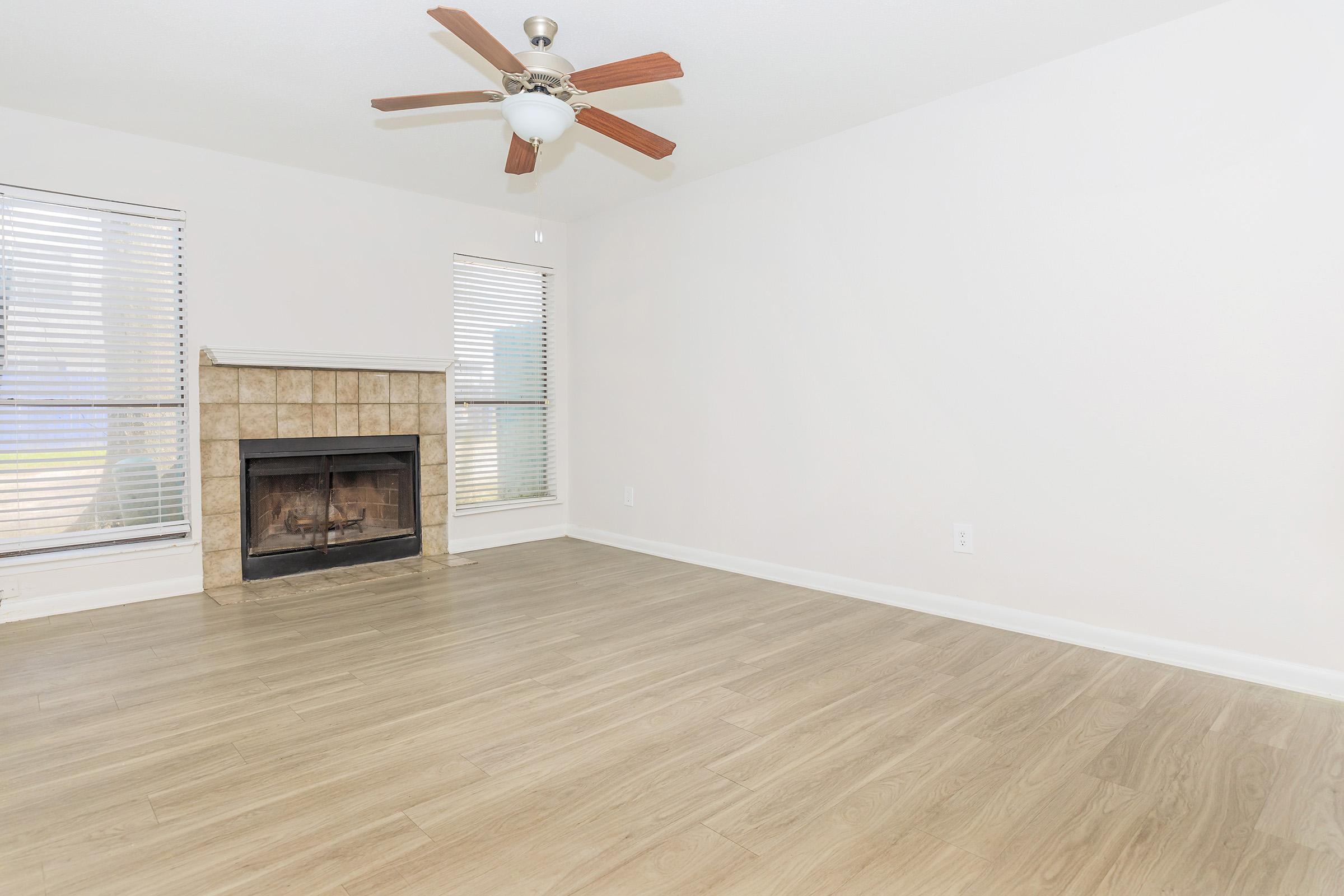 A spacious, empty living room featuring a ceiling fan and a fireplace with a stone surround. Natural light comes through two windows with blinds, and the floor is finished with light-colored wood laminate. The walls are painted in a neutral tone, creating a bright and airy atmosphere.