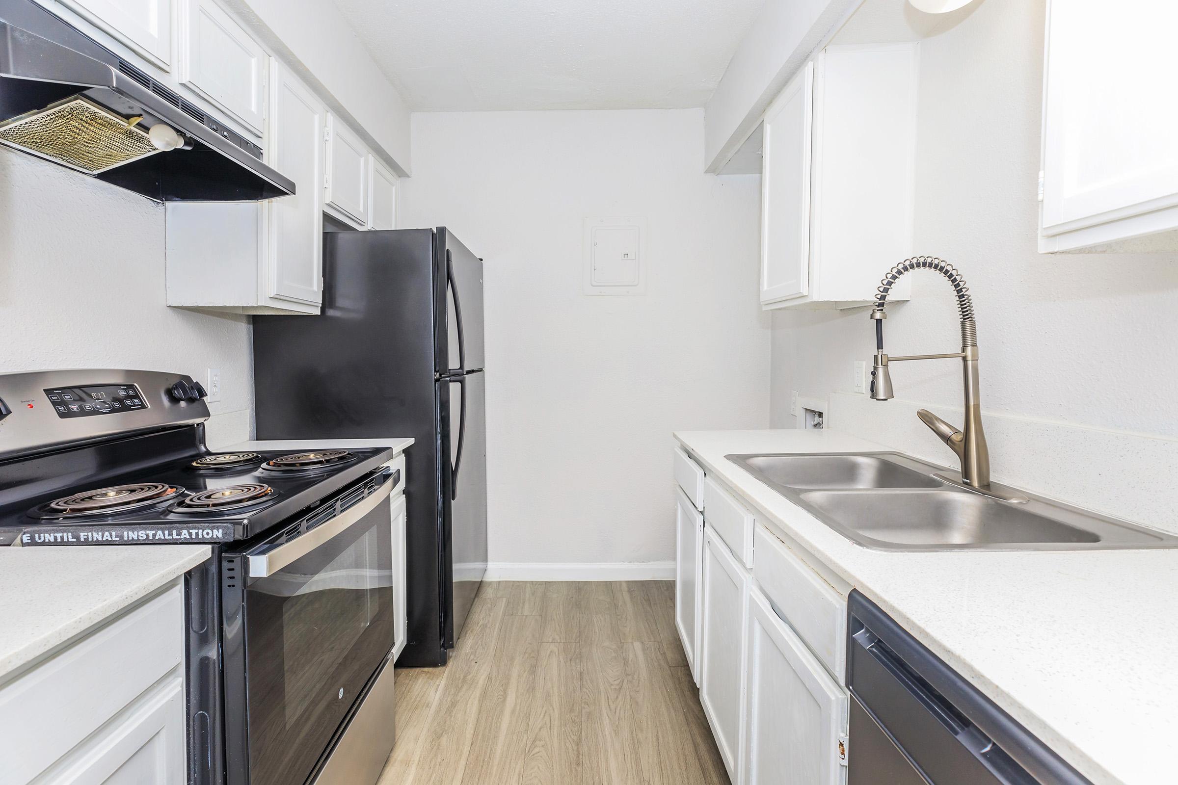 Modern kitchen featuring white cabinets, a dark refrigerator, a stainless steel sink with a faucet, and a black stove with an oven. The countertops are light-colored, and the flooring is wooden. The walls are painted white, creating a bright and spacious atmosphere.