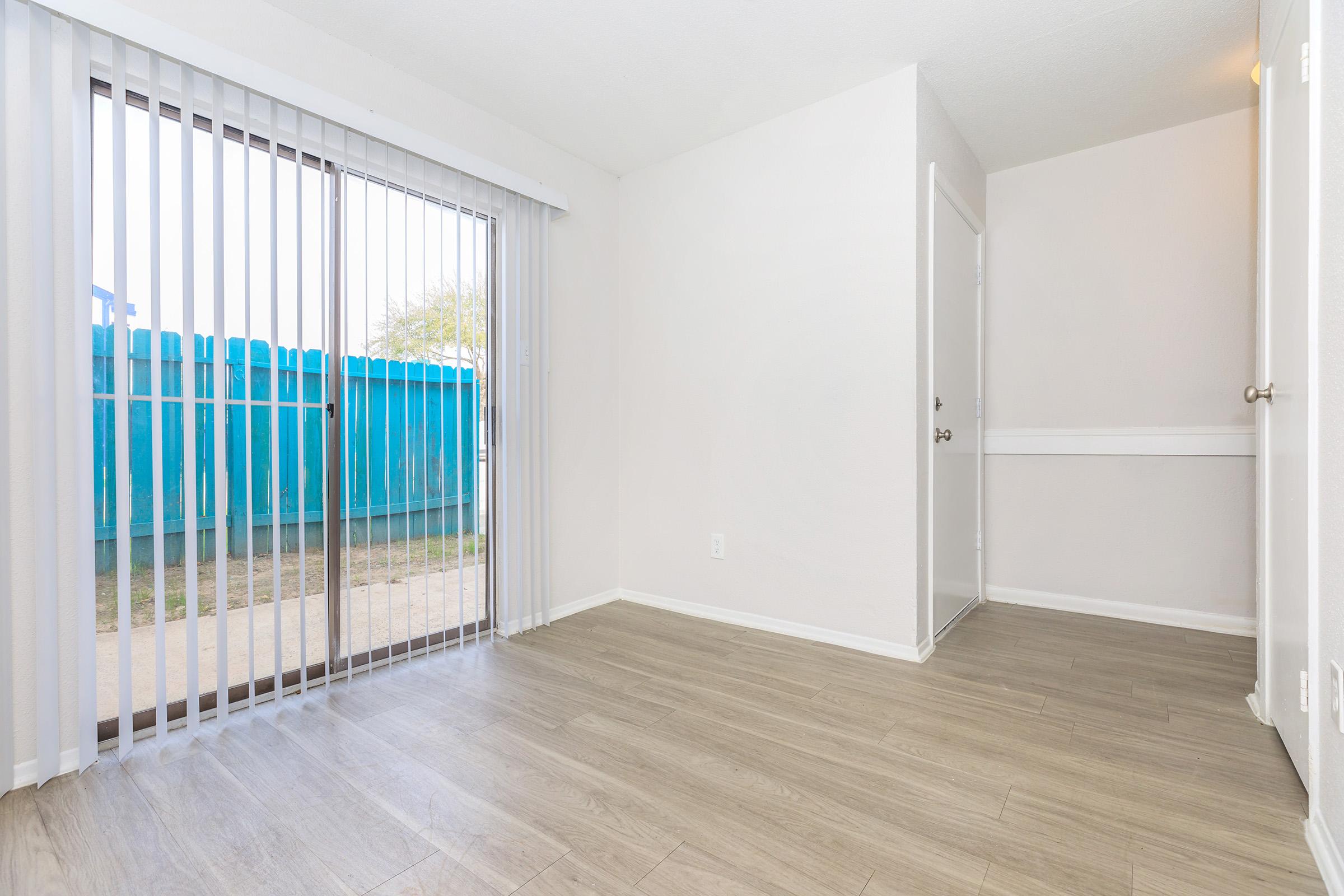 Interior of an empty room with light-colored walls and wood-like flooring. A sliding glass door with vertical blinds leads to an outdoor area, visible through the glass. A blue fence is seen outside. There is a door on the right side leading to another space. The room is bright and uncluttered, conveying a sense of openness.