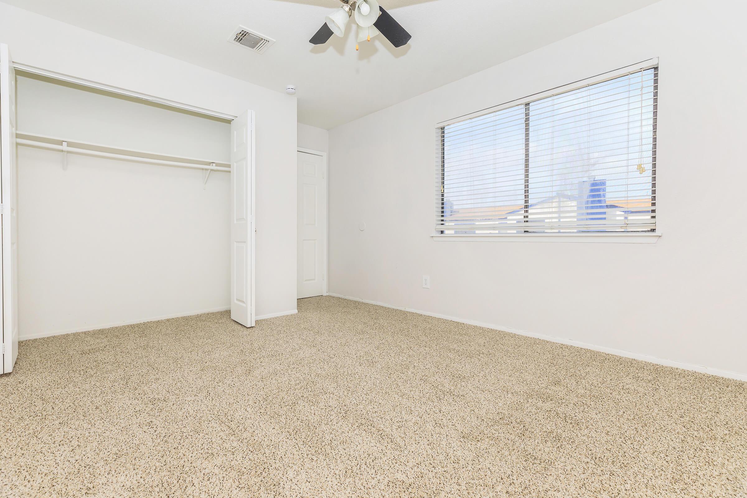 A clean, empty bedroom featuring beige carpet, a single window with horizontal blinds letting in natural light, and a closet with double doors. The walls are painted white, and the ceiling has a ceiling fan. The room is spacious and invites a bright, airy atmosphere.