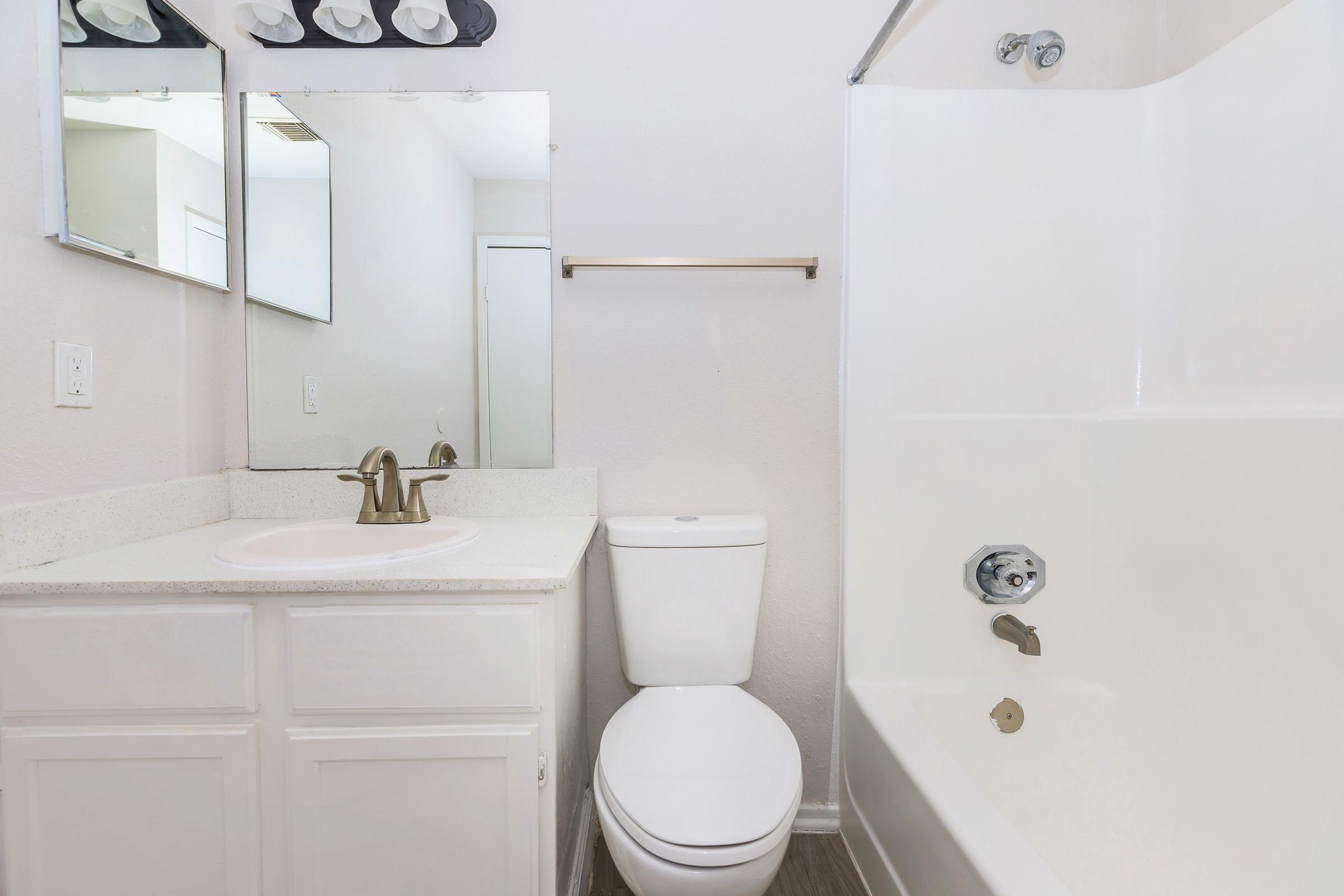 A clean, modern bathroom featuring a white bathtub with a shower, a white toilet, a sink with a beige countertop, and two mirrors above the sink. Soft lighting is provided by a fixture with four bulbs. The walls are light-colored, and there's a towel rack above the toilet.
