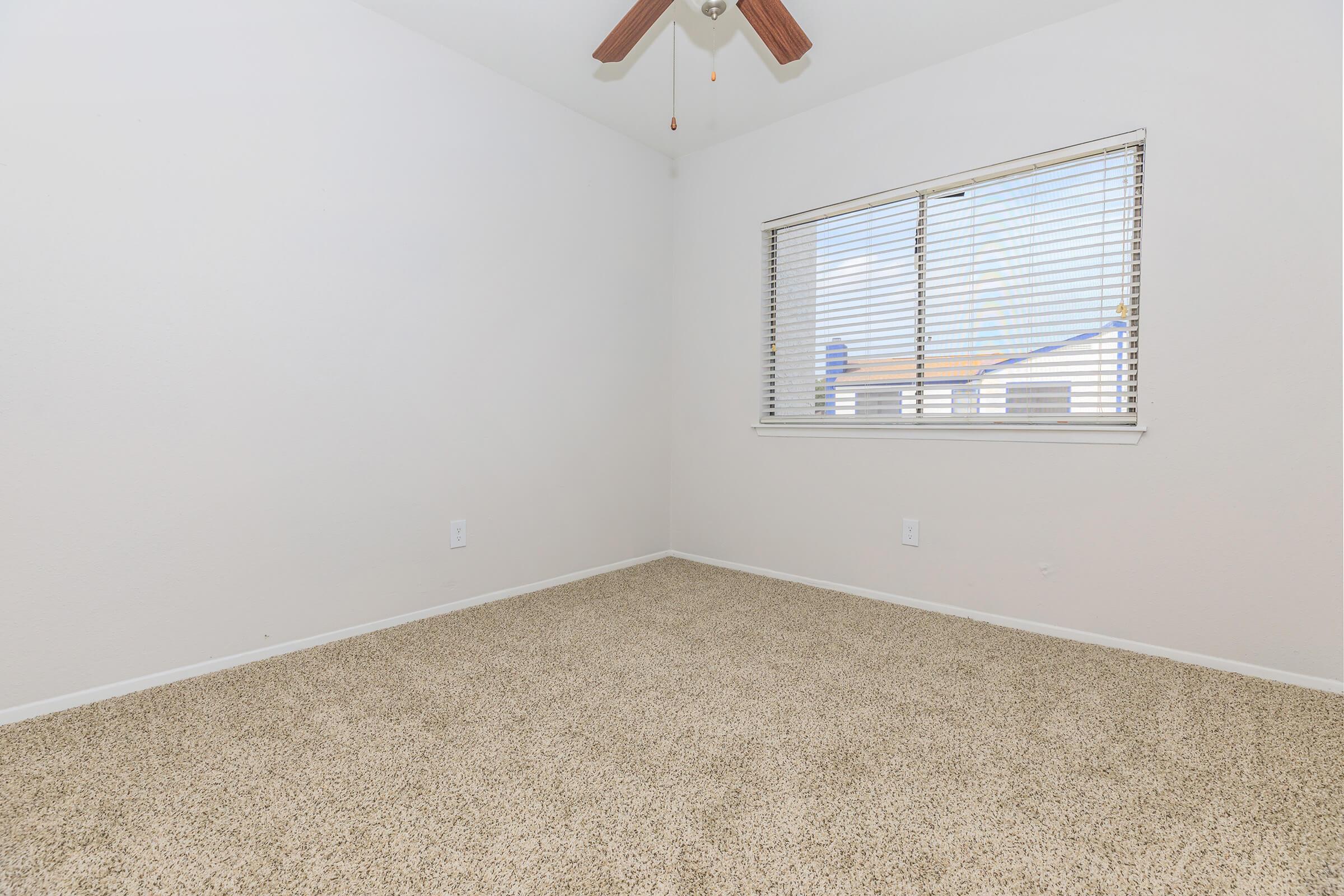 Empty room with beige carpet and white walls. A window with blinds lets in natural light, casting shadows on the floor. The ceiling features a ceiling fan with wooden blades, adding a touch of warmth to the space. The overall ambiance is clean and simple, making it suitable for various purposes.