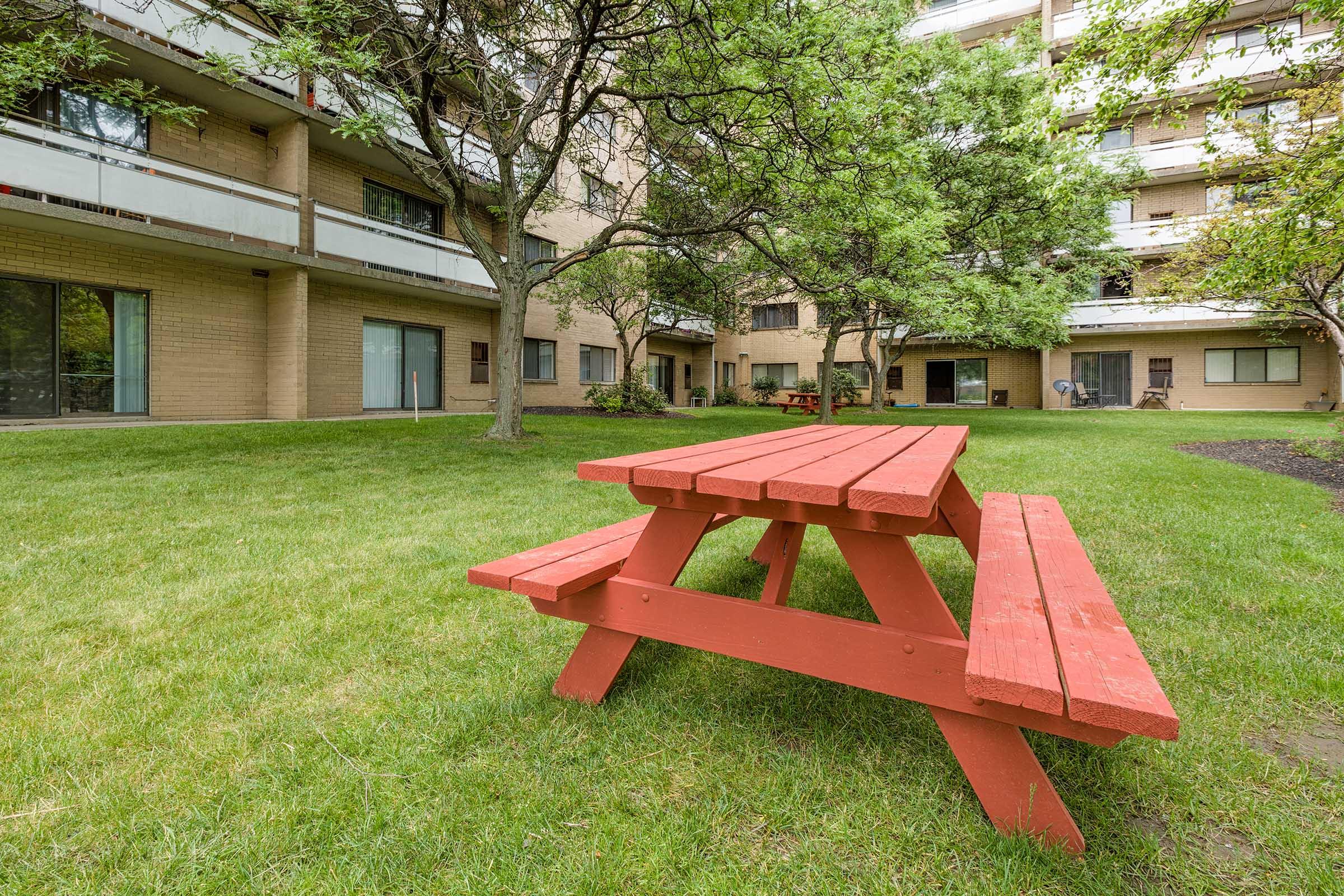 A red picnic table sits on a grassy area surrounded by trees, with several buildings in the background featuring balconies. The scene is part of a courtyard, creating a peaceful outdoor space for relaxation or gatherings.