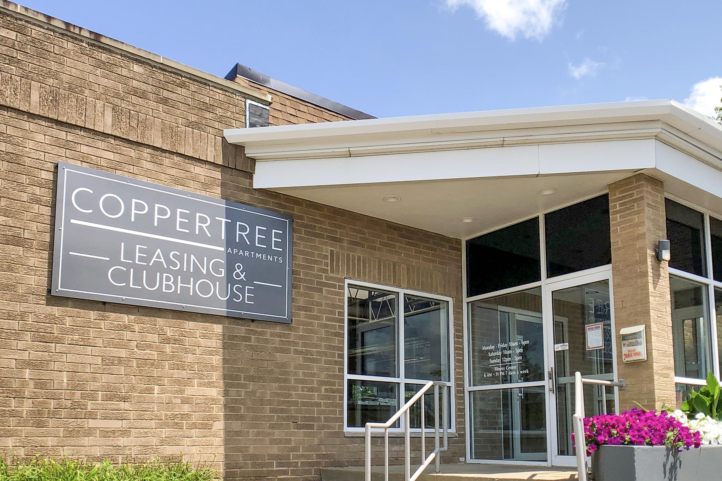 A well-lit entrance of the Copper Tree Apartments leasing and clubhouse building, featuring a sign with the name prominently displayed. The exterior is made of brown bricks, with large windows and a welcoming pathway leading to the entrance, alongside colorful flower arrangements near the entrance.