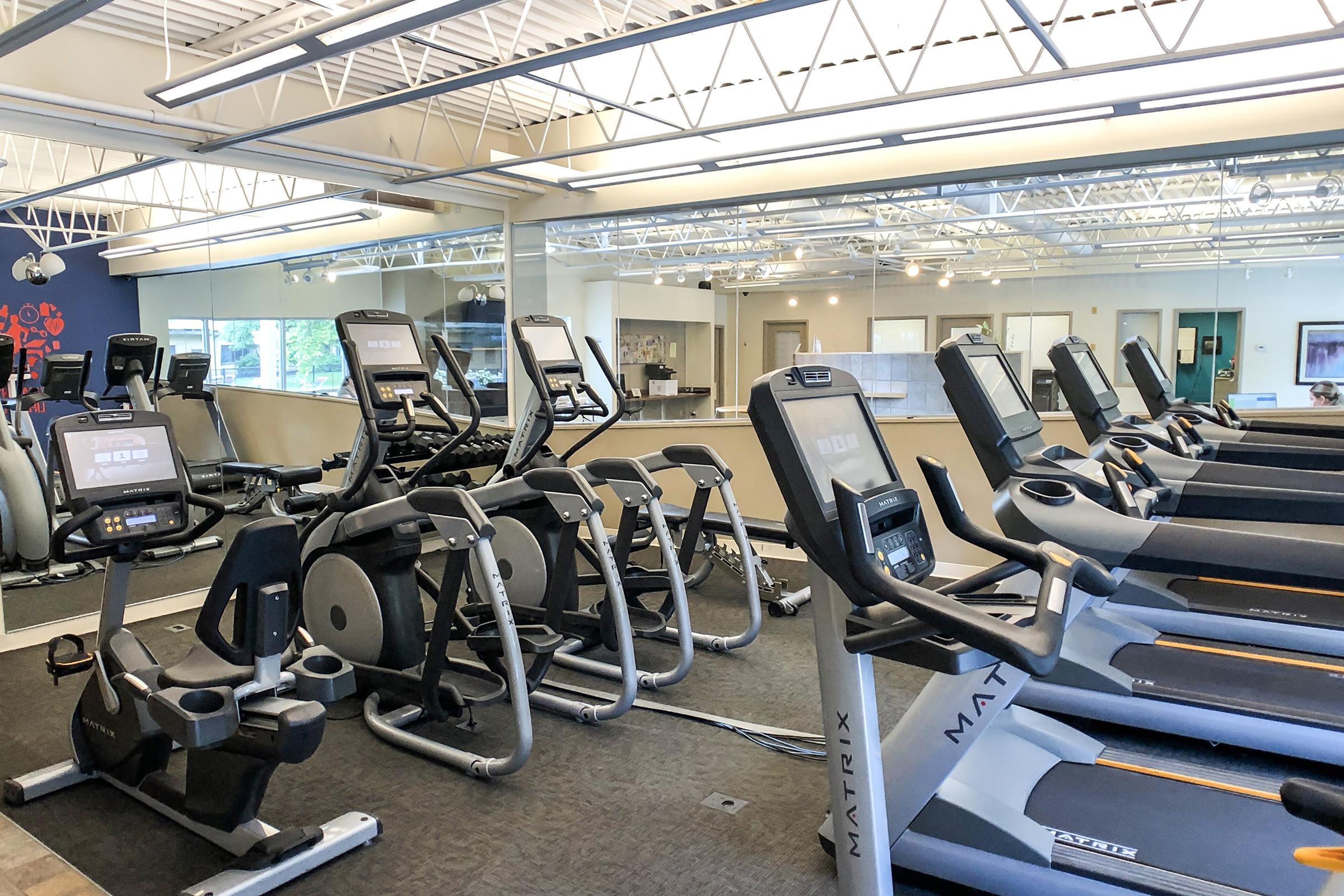 A spacious gym interior featuring various exercise equipment, including treadmills and stationary bikes, arranged neatly. Large windows provide natural light, and mirrors line the walls, enhancing the open atmosphere. The floor is covered in dark carpet, and the overall design is modern and inviting.