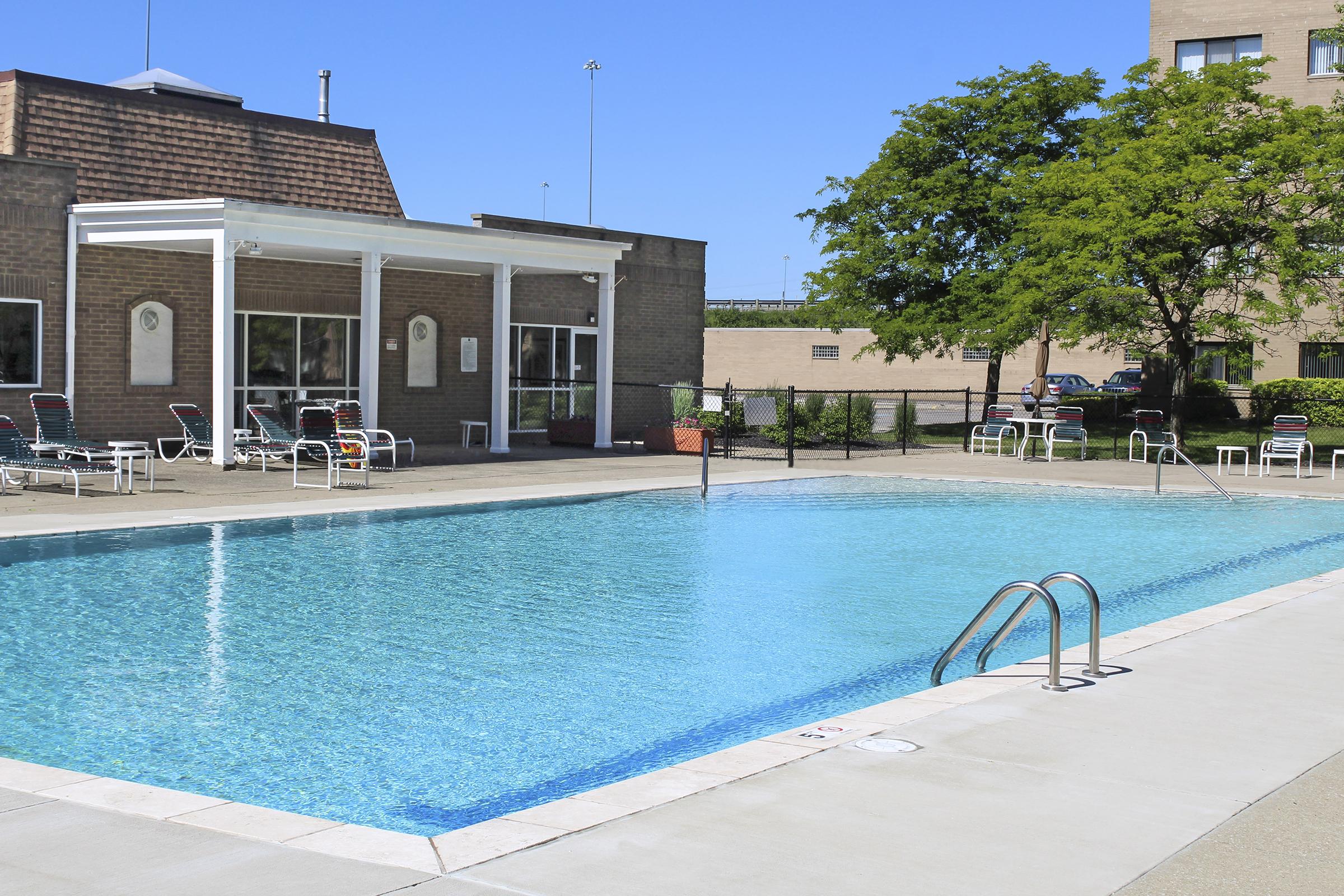 A sunny outdoor swimming pool with clear blue water, surrounded by lounge chairs and a well-maintained area. A building is visible in the background, along with green trees and a clear blue sky. The scene evokes a relaxed atmosphere ideal for leisure and recreation.