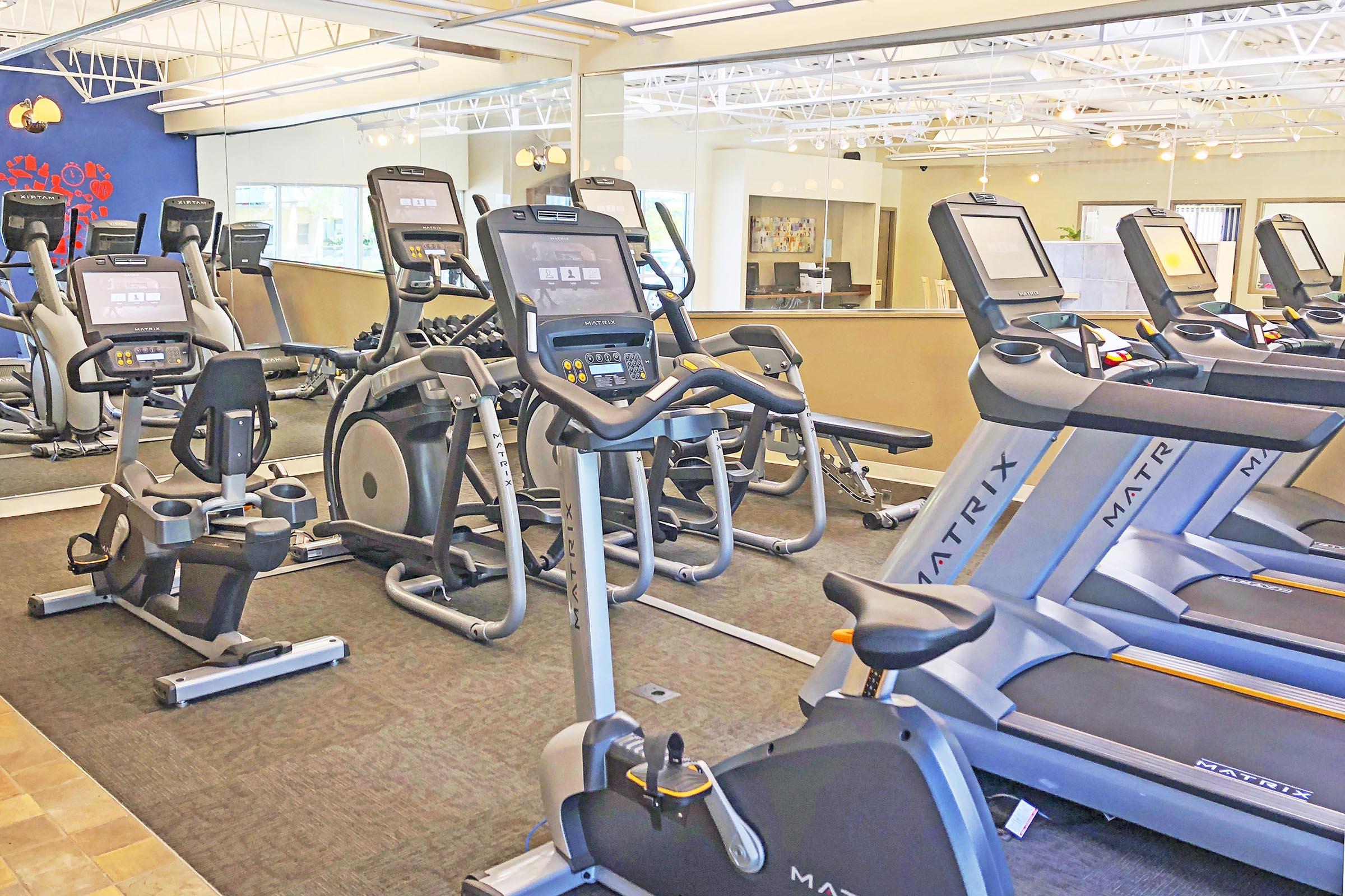 A well-lit gym interior featuring multiple Matrix exercise machines, including treadmills, stationary bikes, and elliptical trainers. The space is organized with carpets and large windows, creating a welcoming atmosphere for fitness enthusiasts.
