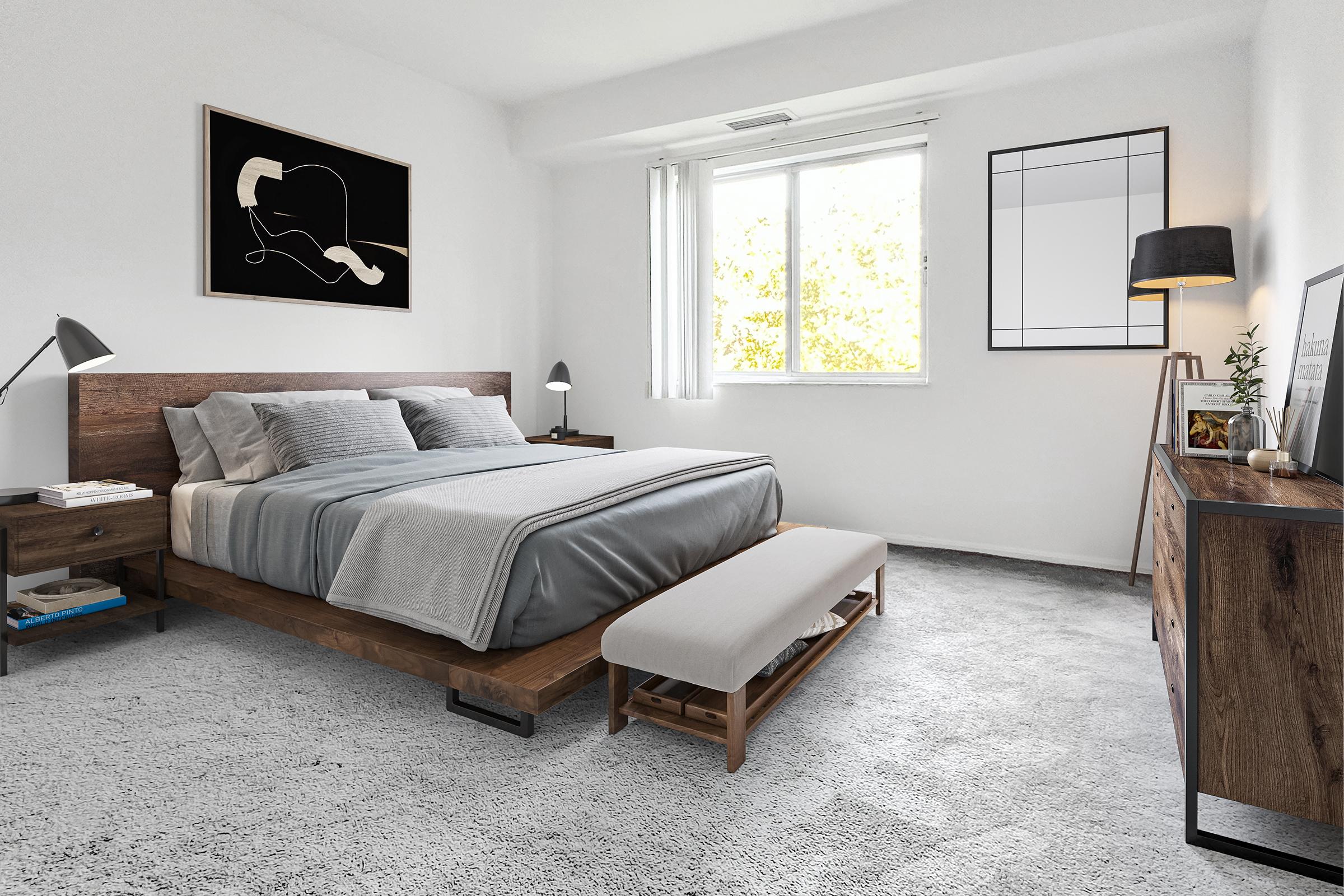 A modern bedroom featuring a wooden bed frame with gray bedding, two bedside lamps, a minimalist painting on the wall, a light-colored rug, a bench at the foot of the bed, a dresser, and a large window allowing natural light in. The overall decor is sleek and contemporary.