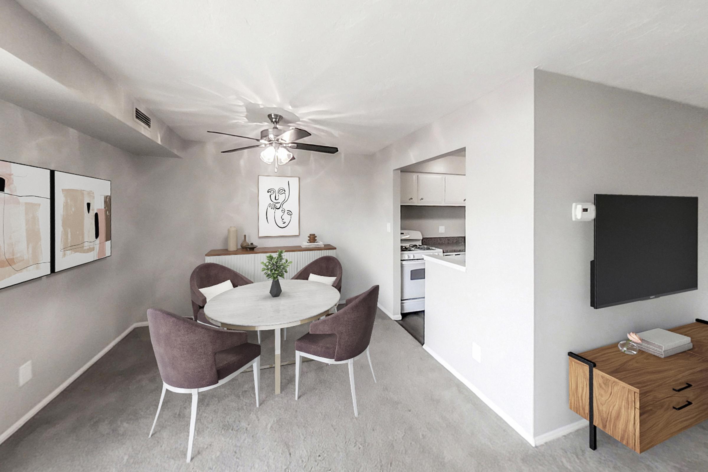 A modern dining area featuring a round table surrounded by four plush chairs. The walls are painted a soft gray, with abstract artwork hanging on one wall. A small kitchen is visible in the background, and there’s a ceiling fan above the table. A stylish wooden media console with a TV is also present in the room.