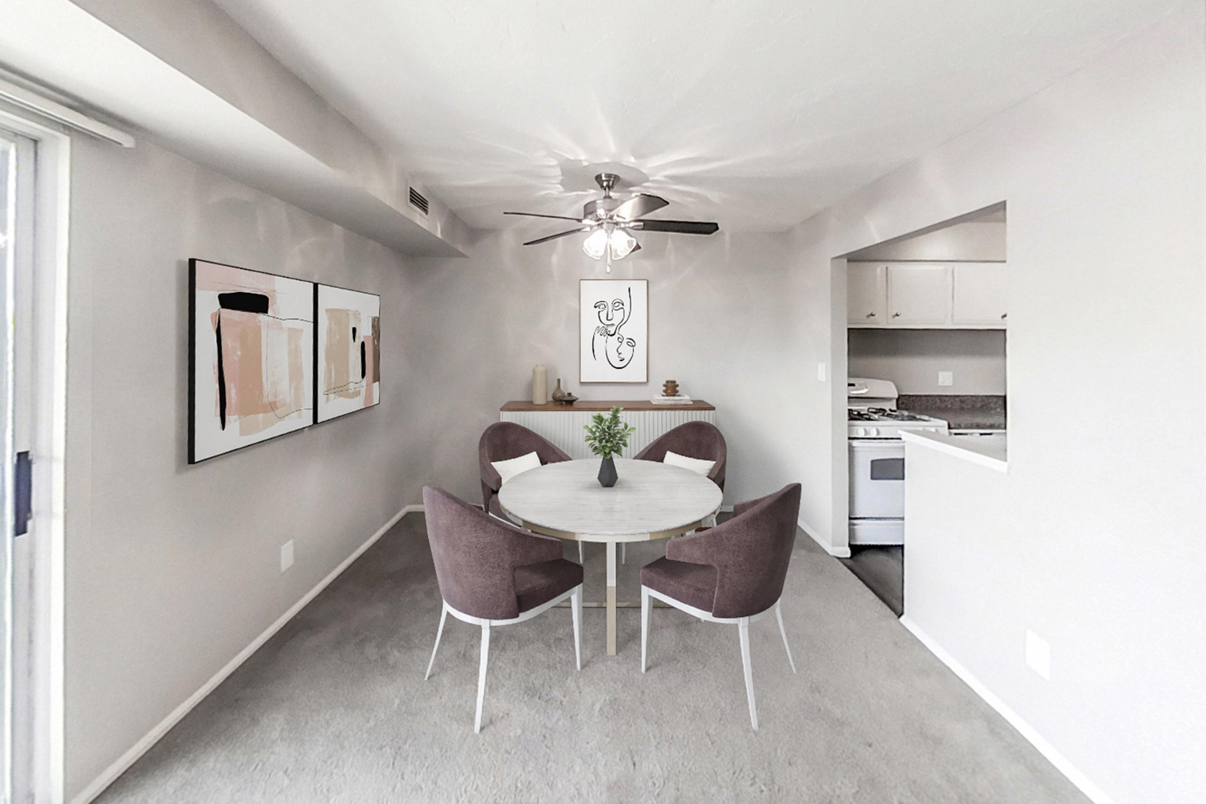 A modern dining area featuring a round table with four upholstered chairs. Two abstract art pieces adorn the wall. A ceiling fan hangs above, and the adjacent kitchen is visible with white cabinets and a stove. The walls are a soft gray, creating a cozy atmosphere.