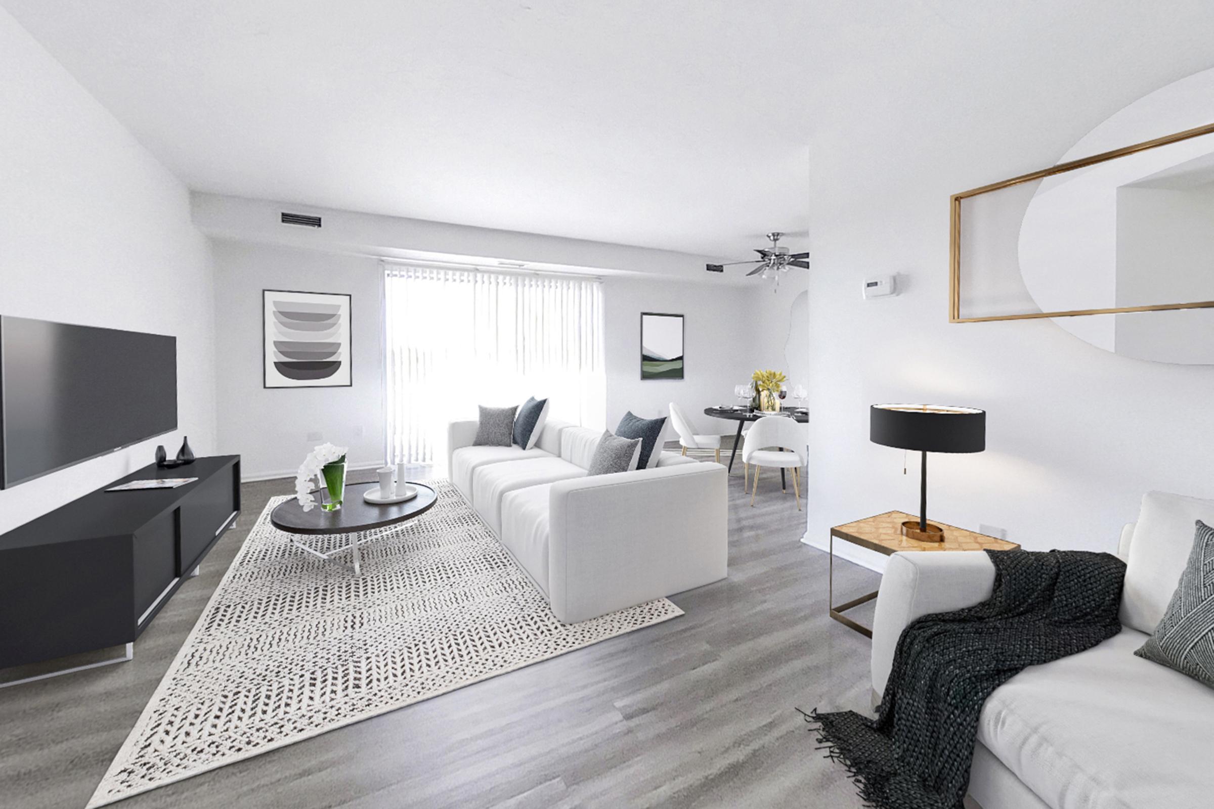 A modern living room featuring white sofas, a round coffee table, and a patterned area rug. A large TV is mounted on the wall alongside a mirror. Natural light streams in through vertical blinds, highlighting minimalist artwork on the walls. A dining area with a table and chairs is visible in the background.