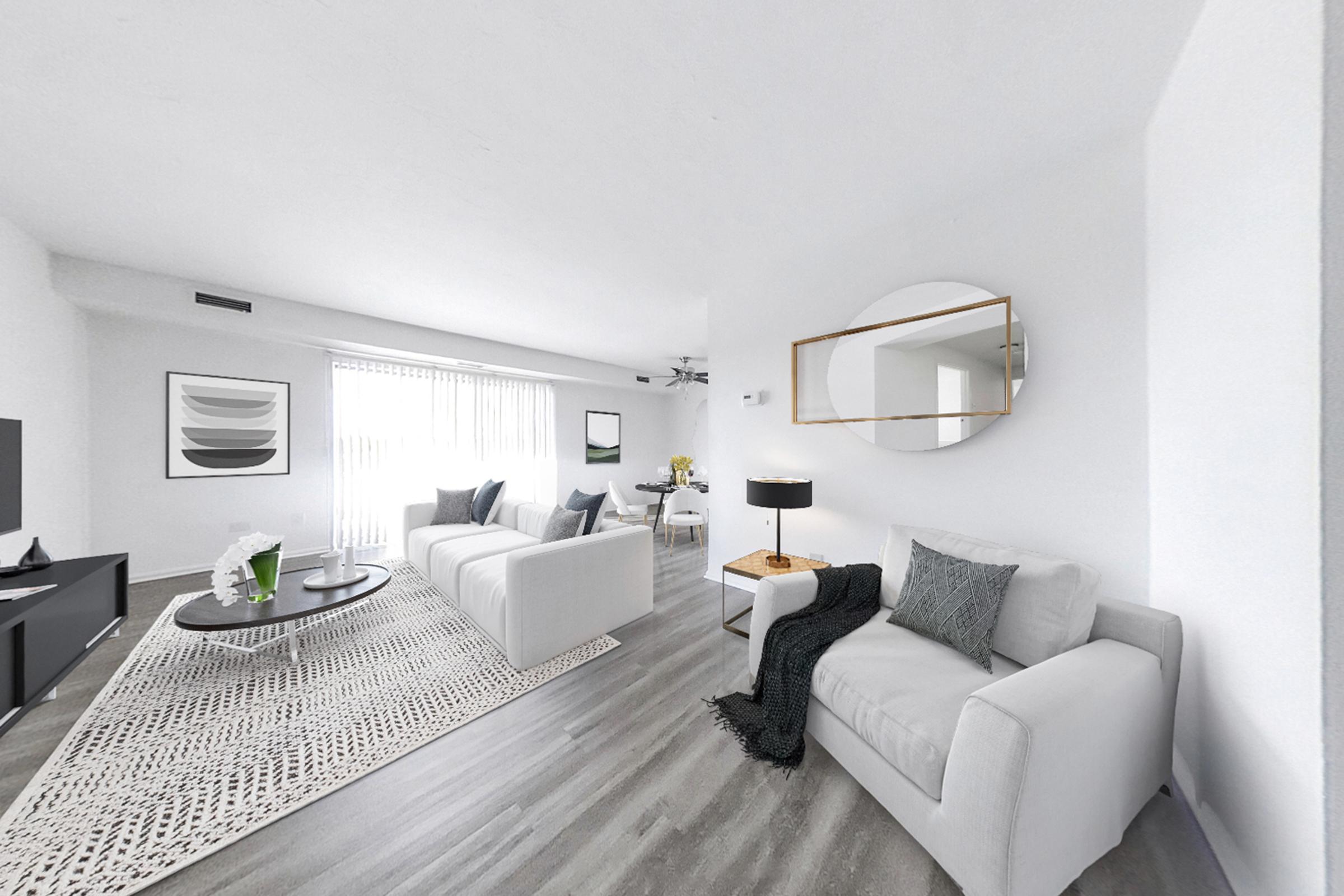 Bright and modern living room featuring two white sofas, a round coffee table, and a stylish area rug. Large windows with sheer curtains allow natural light to fill the space. A wall-mounted mirror and minimalist decor add elegance, while a TV and plants provide a cozy atmosphere.