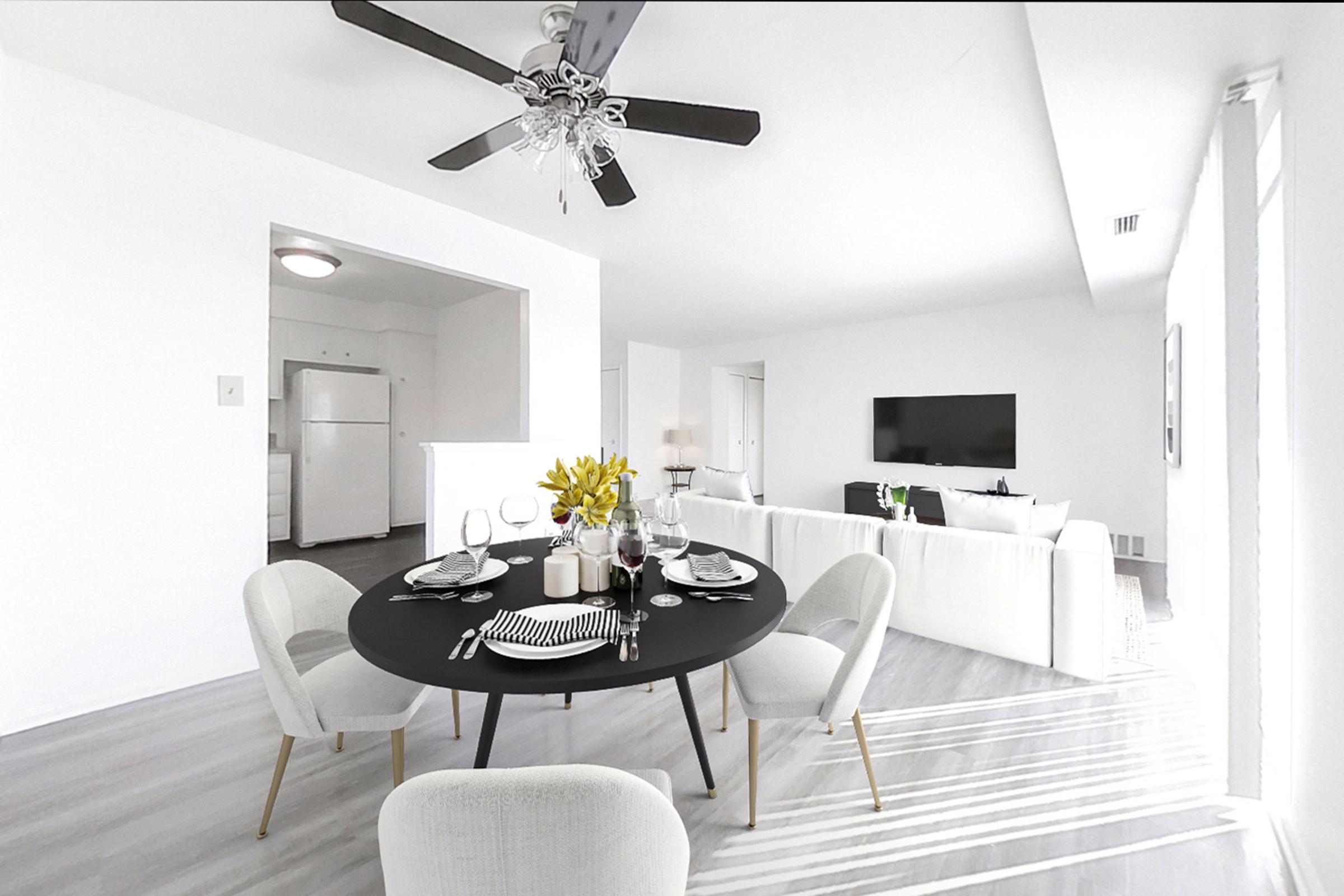 A modern dining area featuring a round black table set with dinnerware and glasses. There are white chairs and a floral centerpiece. In the background, a bright living space includes a white sofa and a television. The room is well-lit, with natural light streaming through large windows.