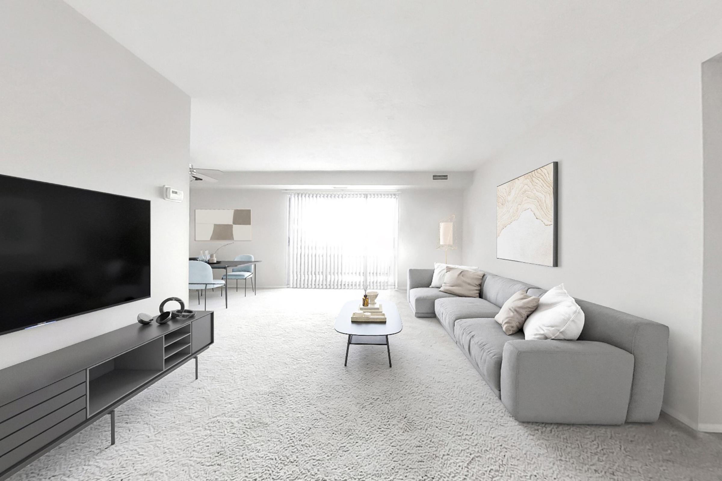 A modern, minimalist living room featuring a light gray sofa with decorative pillows, a sleek coffee table, and a dark media console with a flat-screen TV. Large windows allow natural light to fill the space, adorned with white vertical blinds. A dining area with a round table and chairs is visible in the background.