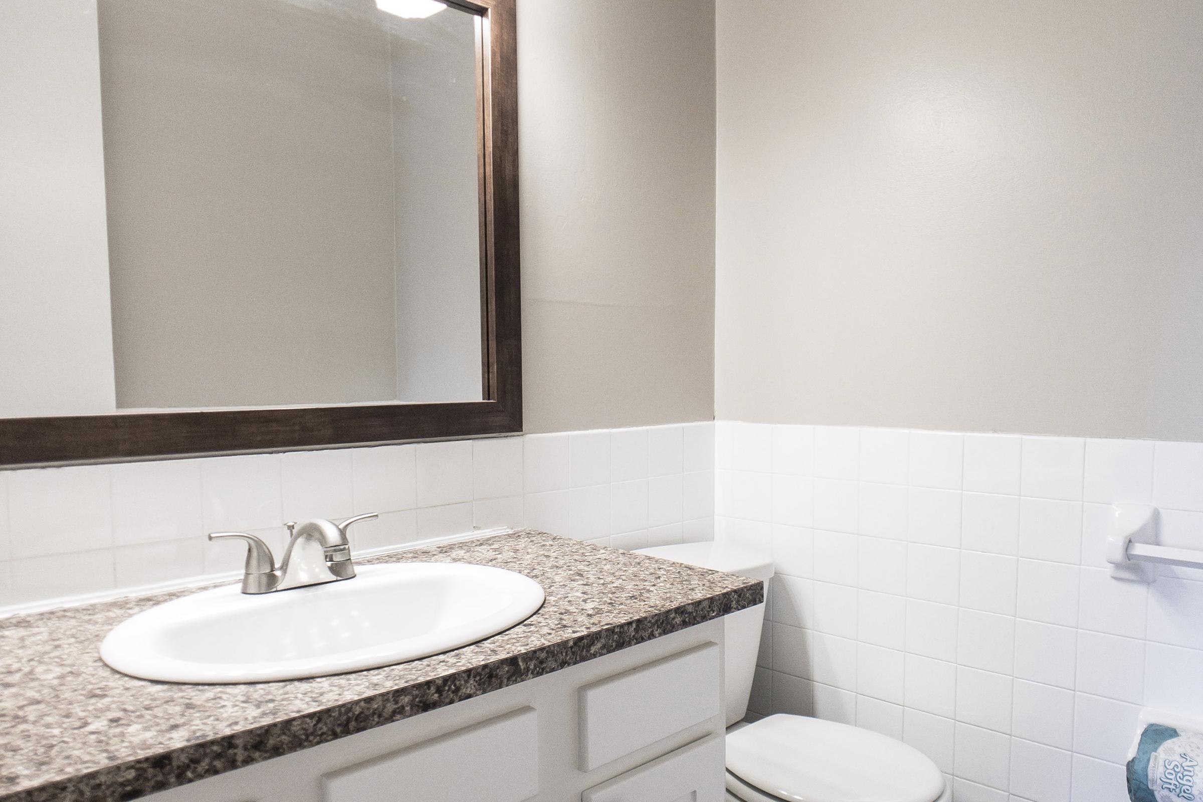 A clean and modern bathroom featuring a white sink with a granite countertop, a large mirror above the sink, a white toilet, and beige walls. The tiled wall adds a stylish touch, while a towel is hanging nearby, completing the simple yet elegant decor.