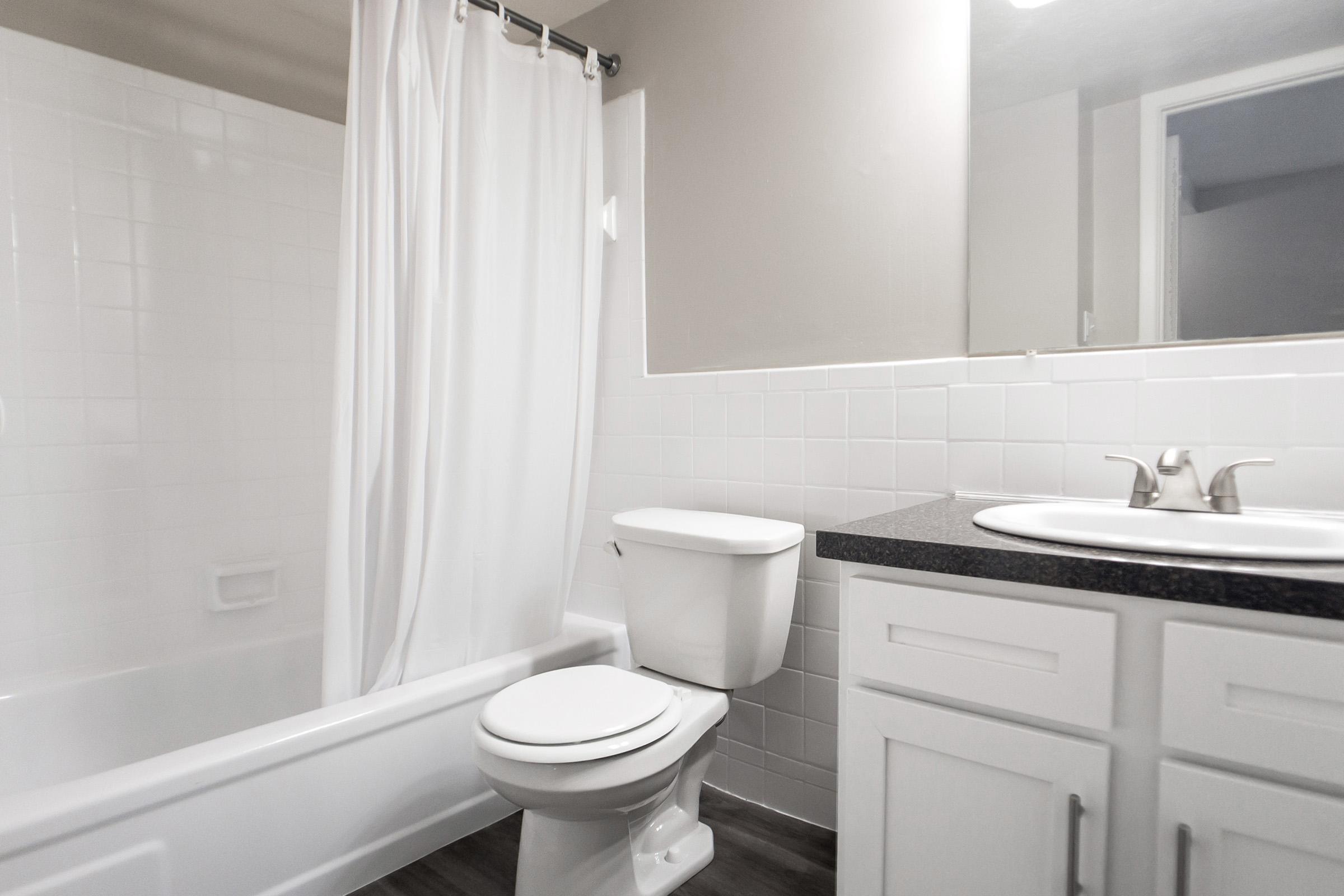 A small, modern bathroom featuring a bathtub with a white shower curtain, a white toilet, and a sink with a black countertop. The walls are tiled in white, and there is a mirror above the sink. The floor appears to have a dark wood-like finish.