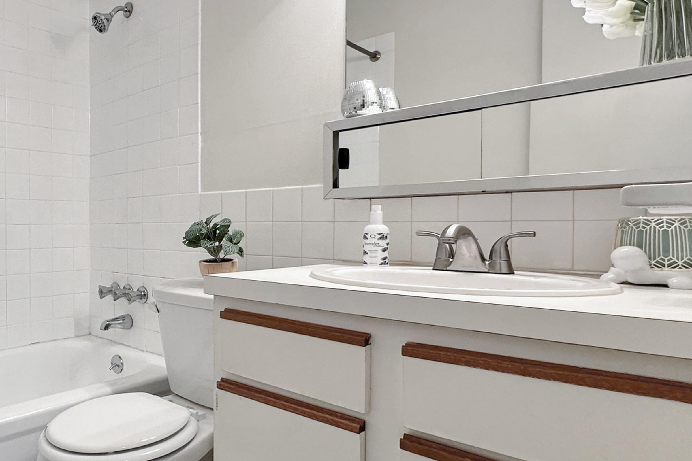 A clean and modern bathroom featuring a white vanity with wooden accents, a sink, a toilet, and a bathtub. The mirror above the sink reflects a bright space, and there's a small potted plant on the countertop. Tiled walls and neutral colors create a fresh, minimalist atmosphere.