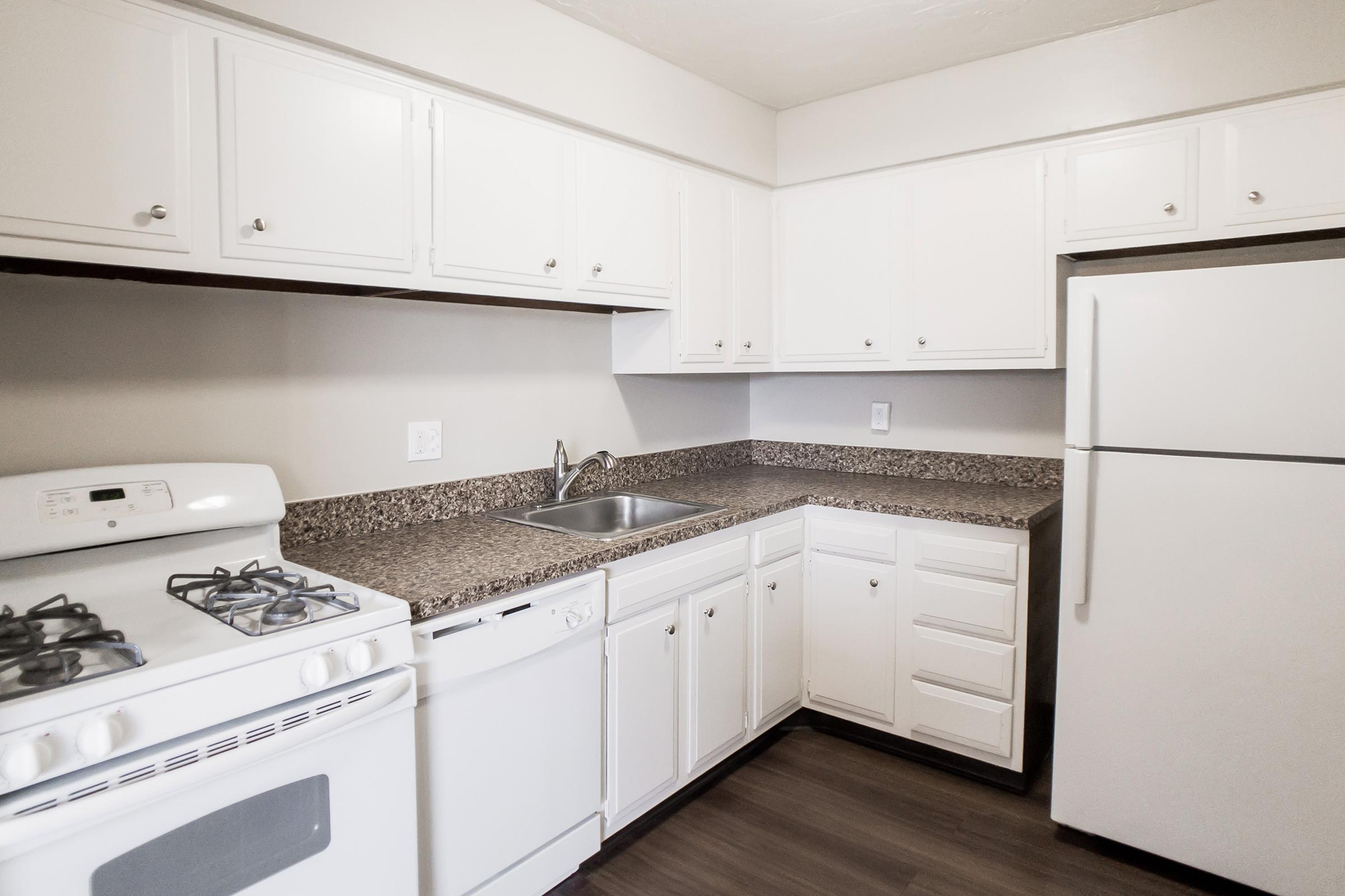 A modern kitchen featuring white cabinetry, a stainless steel sink, and a gas stove. The counter has a brown speckled surface, and there's a side-by-side refrigerator. The space is well-lit and clean, with a simple and functional layout ideal for cooking and meal preparation.