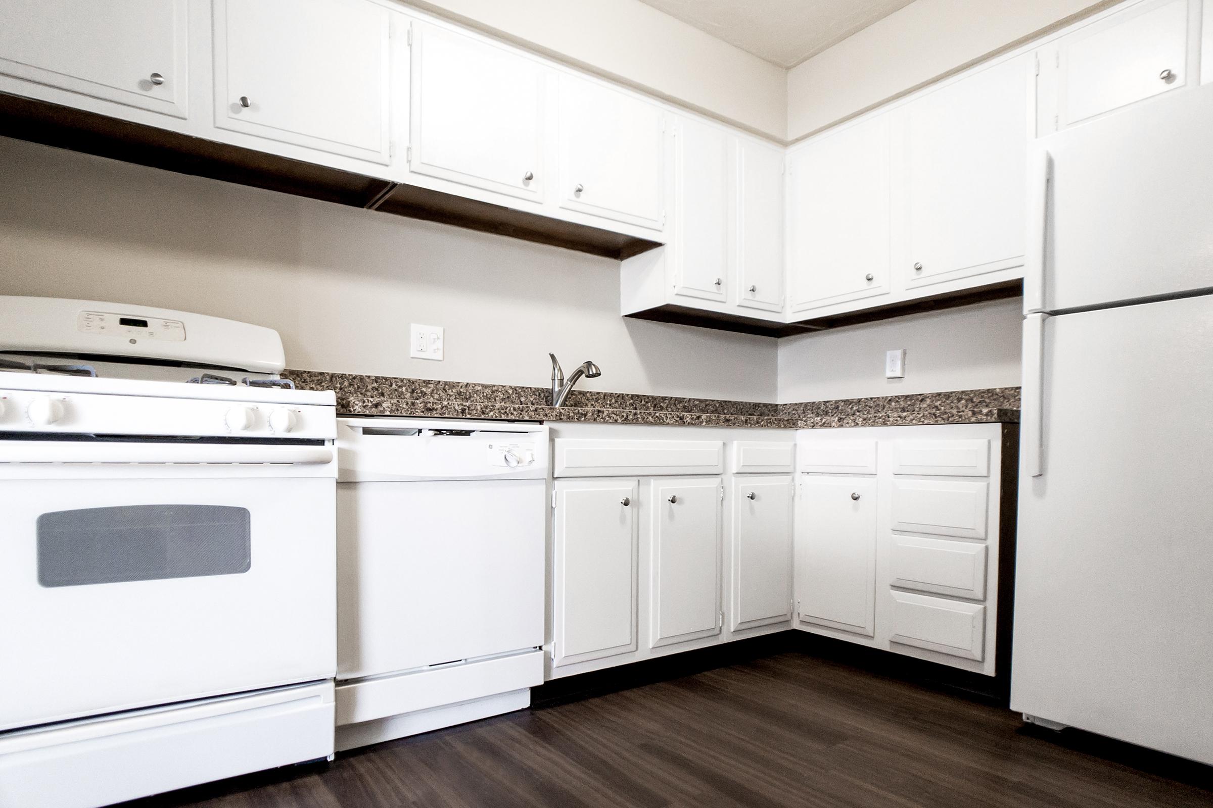 A modern kitchen featuring white cabinetry, a gas stove, a dishwasher, and a refrigerator. The countertops are a speckled granite, and the flooring is a warm wood finish. The kitchen has a clean and organized appearance, with ample storage space.