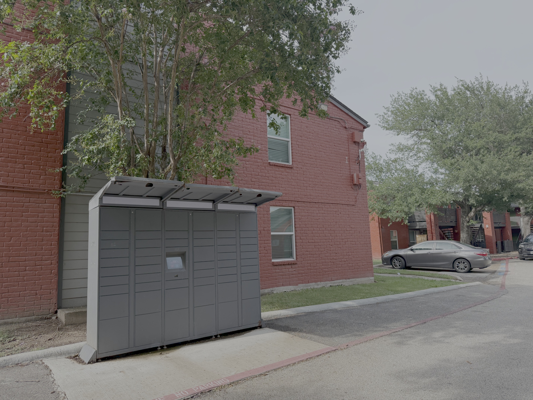 A gray parcel locker unit is positioned next to a red brick apartment building, partially shaded by a small tree. Nearby, there is a parking area with a silver car and additional apartment buildings in the background, surrounded by green grass and trees.