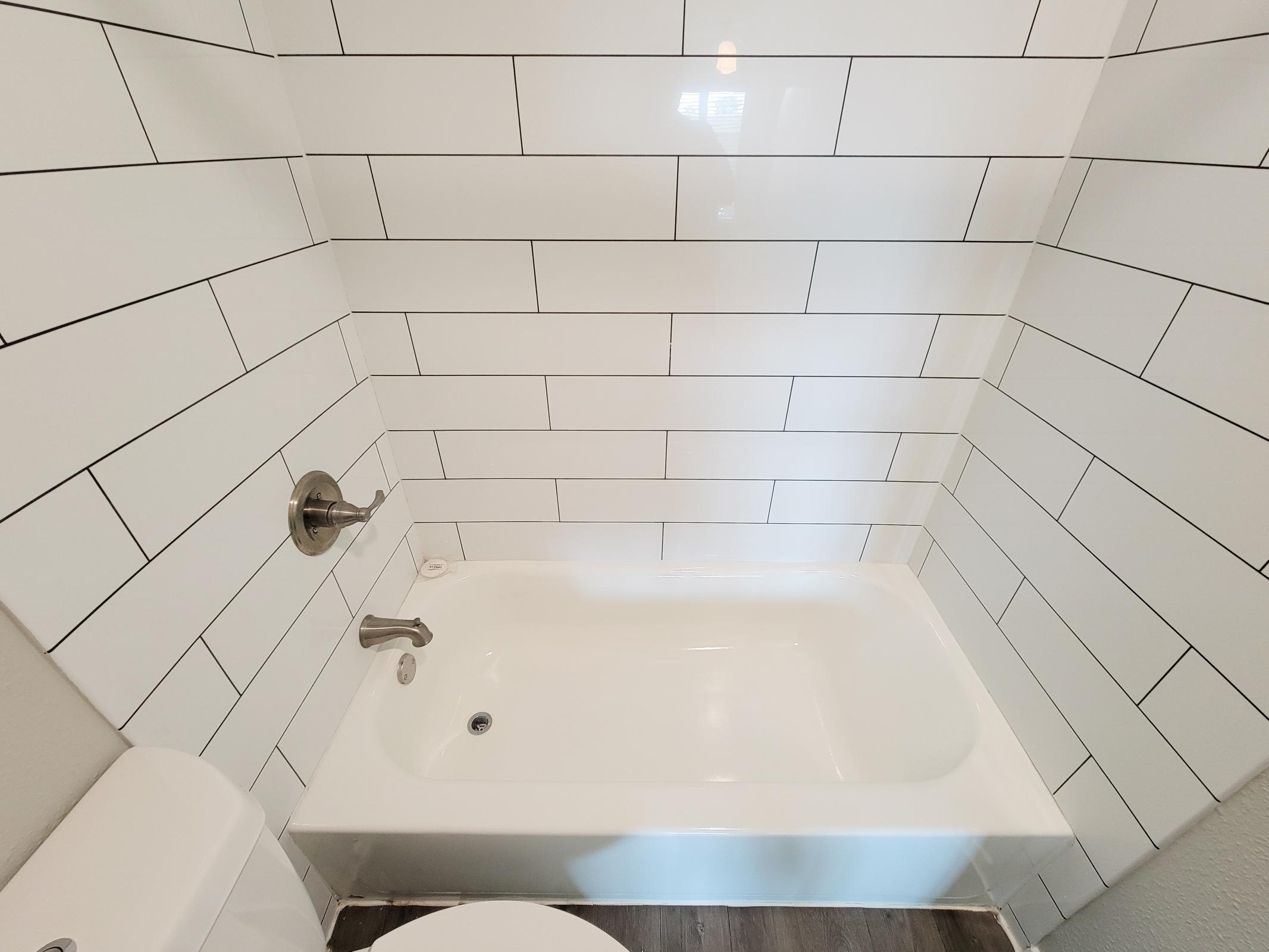 A modern bathroom featuring a white tub with chrome fixtures, surrounded by white tiled walls with black grout lines. The floor is dark wood, adding contrast to the airy space. A portion of a toilet is visible on the left side of the image. Natural light is reflected on the tiles.