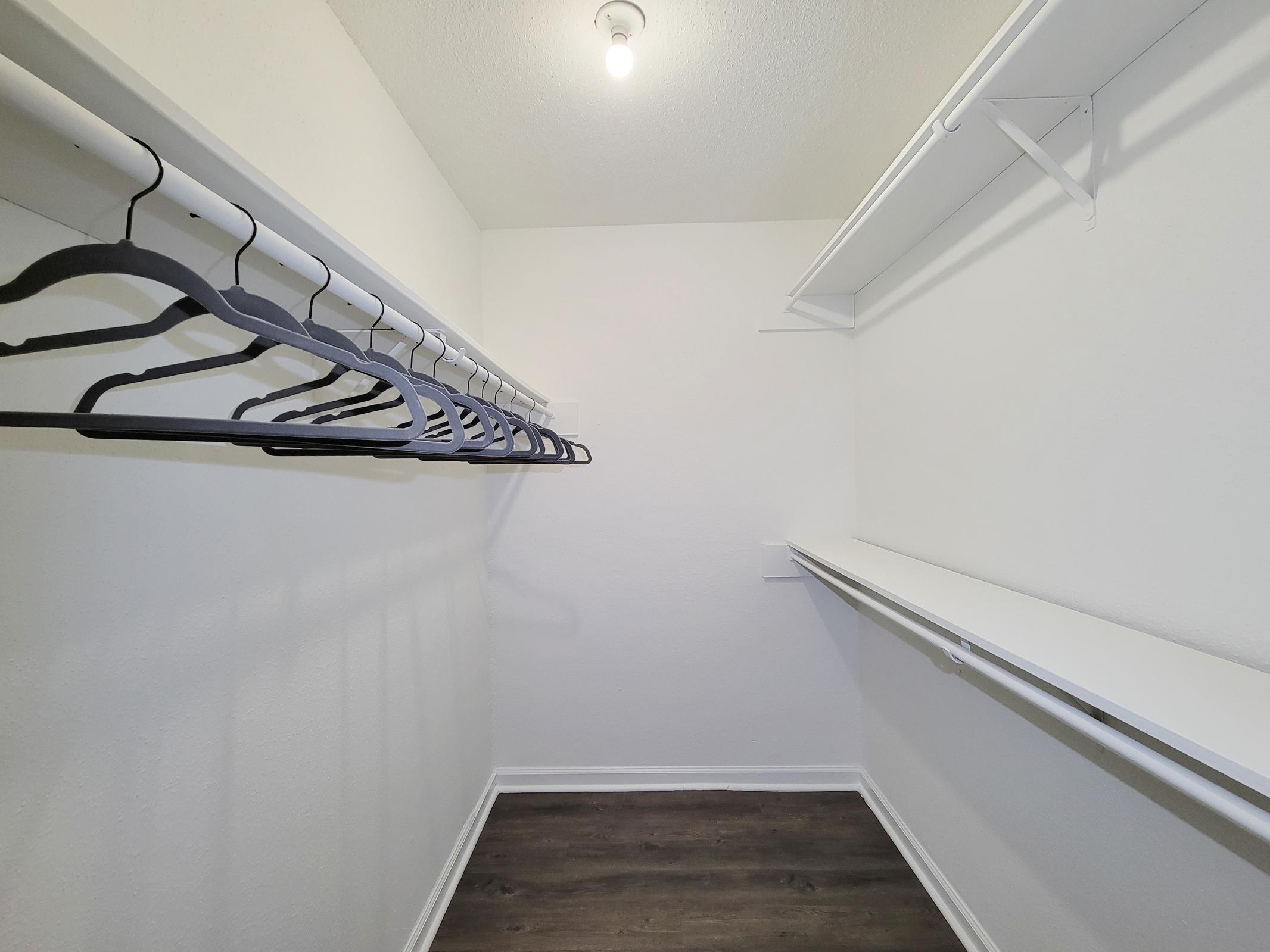 A clean and spacious walk-in closet featuring white walls and a light fixture. The closet has multiple hanging rods with black hangers and a long shelf along one side. The flooring is dark wood, adding a modern touch to the minimalistic design.