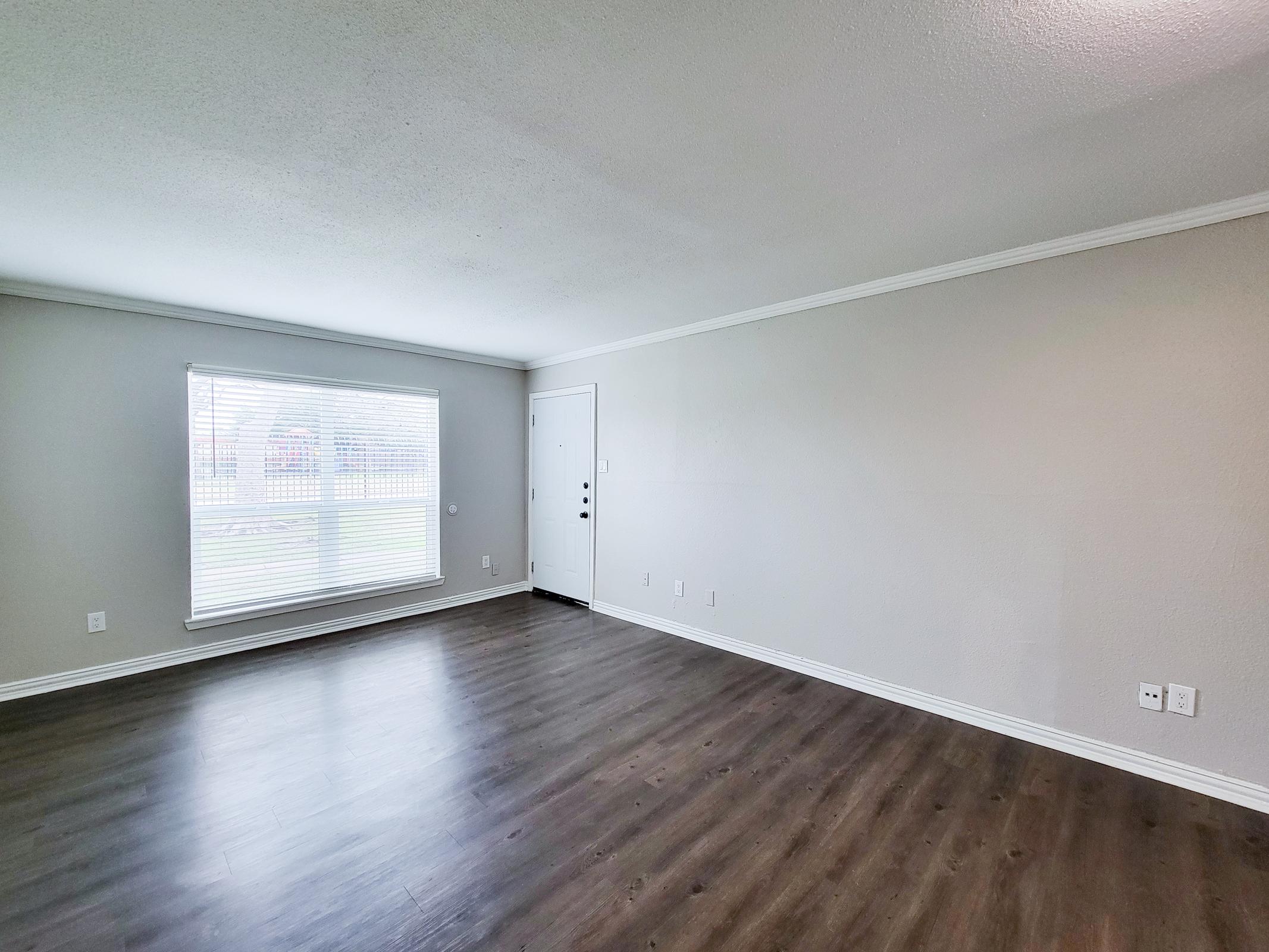 A spacious living room featuring light gray walls, a large window letting in natural light, and a front door. The flooring is dark wood, and there are minimal decorations or furniture, creating a clean and open atmosphere.