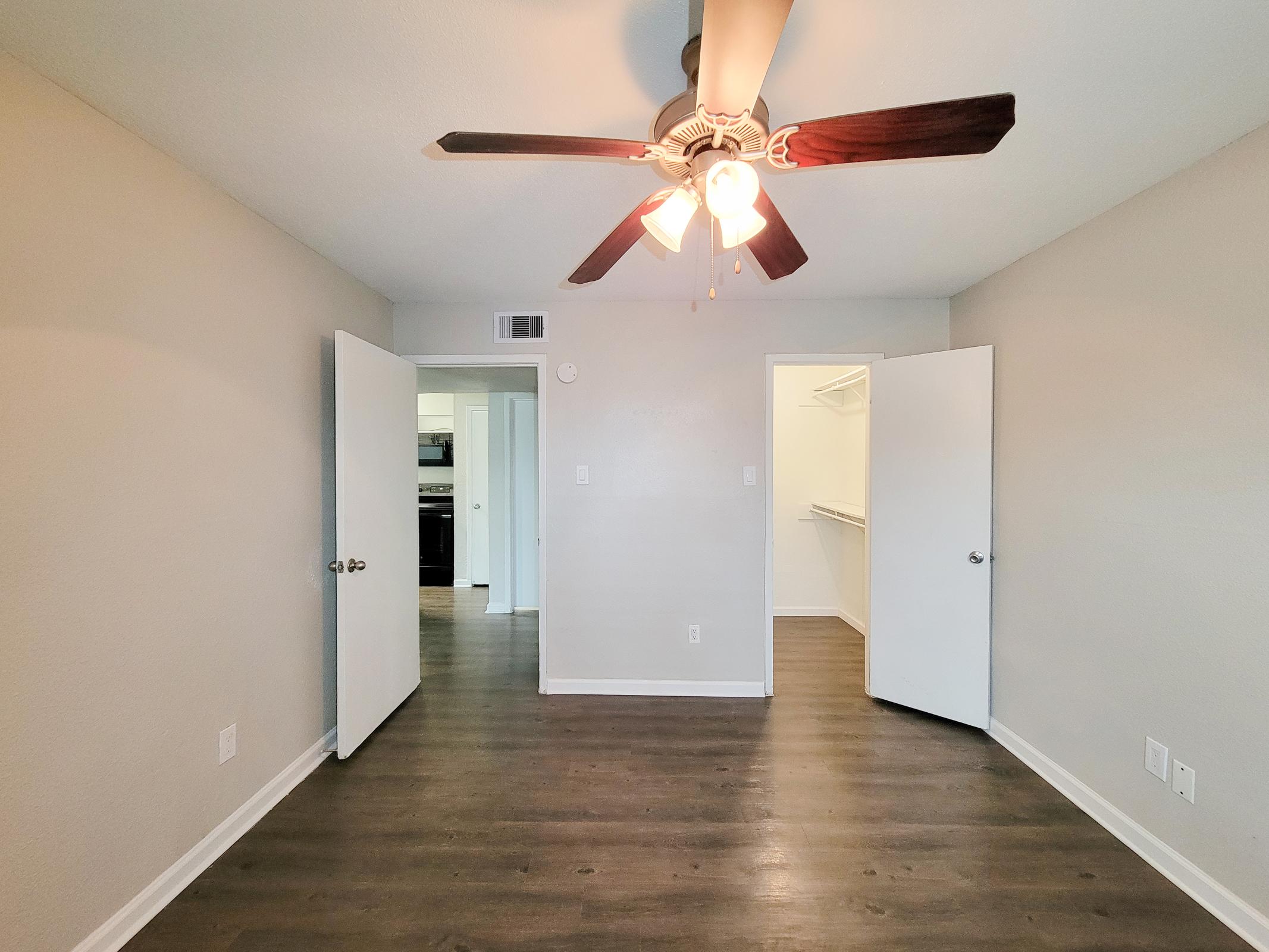 A spacious, well-lit room featuring a ceiling fan with three light bulbs. Two white doors are open to reveal a closet on one side and an entrance to another room on the other. The walls are painted a neutral color, and the floor is covered in dark wood laminate.