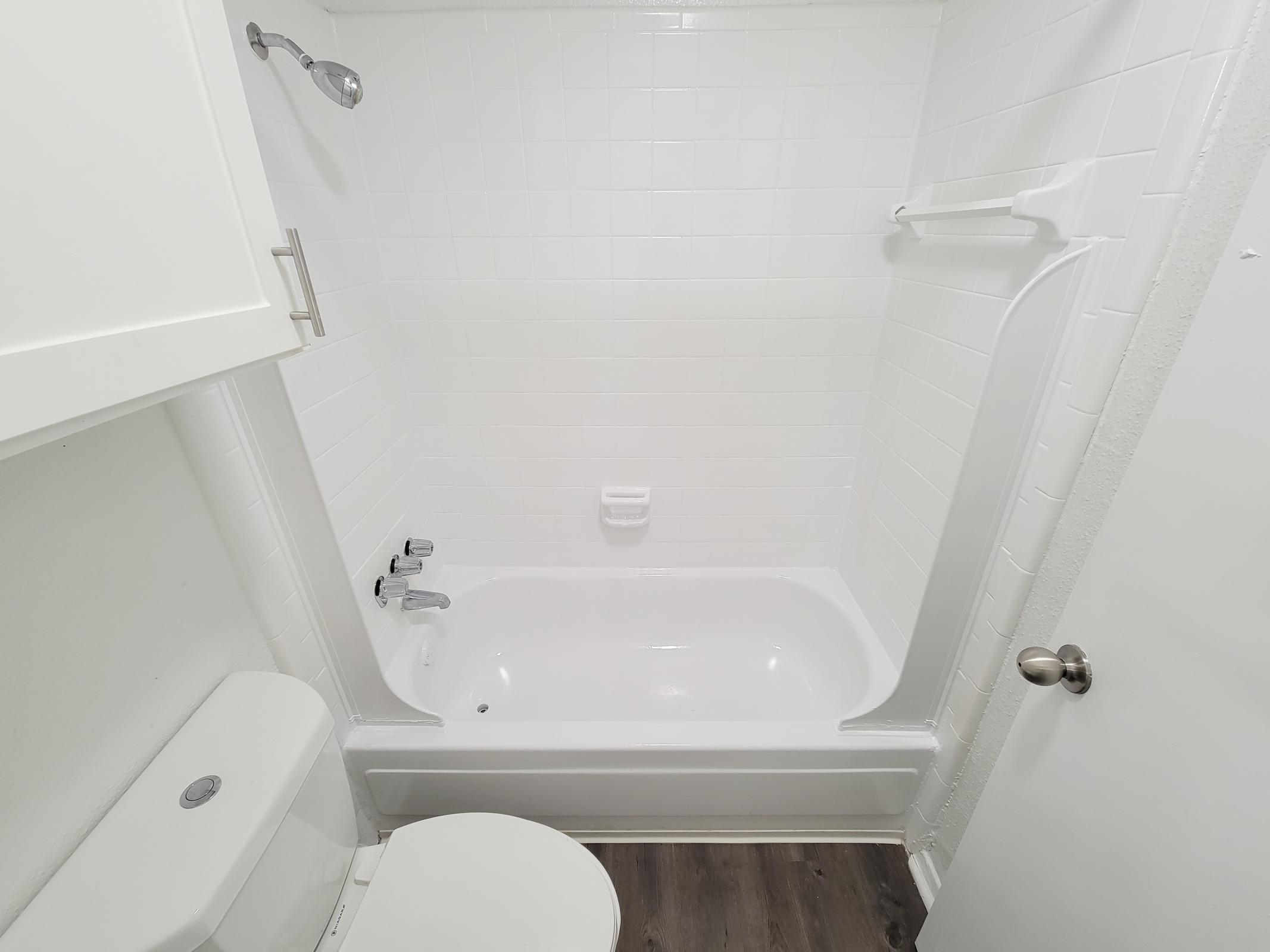 A clean, modern bathroom featuring a white tub and shower combination, a toilet, and light-colored walls. The floor has a wood-like finish, and there is a towel bar and a door leading outside the bathroom. Overall, the space is bright and minimalistic.