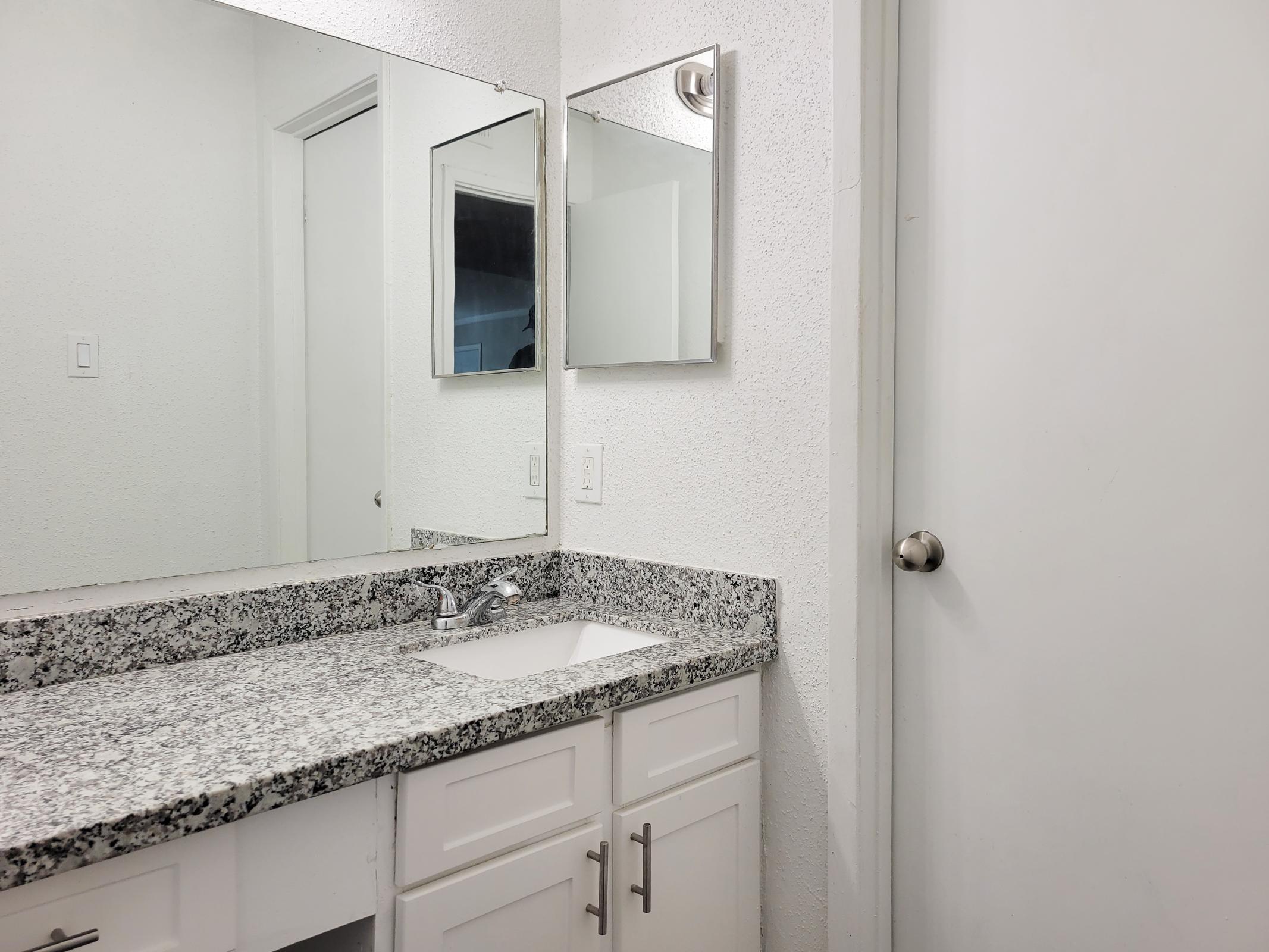 A clean, modern bathroom featuring a granite countertop with a sink, a large mirror above, and white cabinetry. A light-colored wall complements the simple design. A closed door is visible on the right, adding to the spacious feel of the room. Natural light enters, enhancing the bright ambiance.