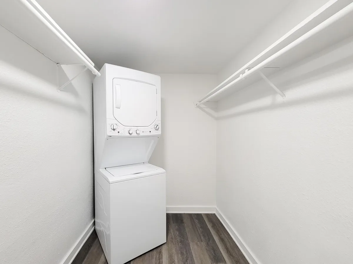 A compact laundry area featuring a stacked washer and dryer in a small, well-lit space. The walls are plain white, and there are two hanging rods on either side for clothing storage. The flooring is a light wood-like laminate.