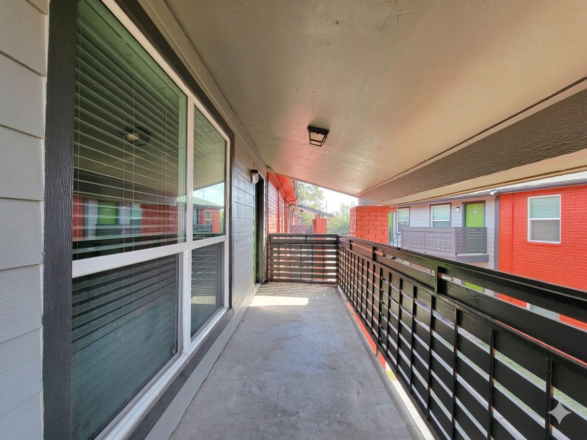 A covered balcony with a railing, decorated with wooden panels and large windows, showing a view of nearby buildings. The setting is well-lit, with sunlight illuminating the light gray concrete floor and the walls of the adjacent apartments in red and cream colors.