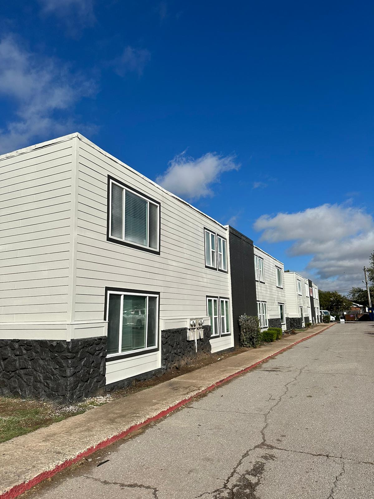 A row of modern, two-story apartment buildings with a mix of white and gray siding. The buildings have large windows and a well-maintained exterior, situated alongside a paved street under a partly cloudy blue sky.