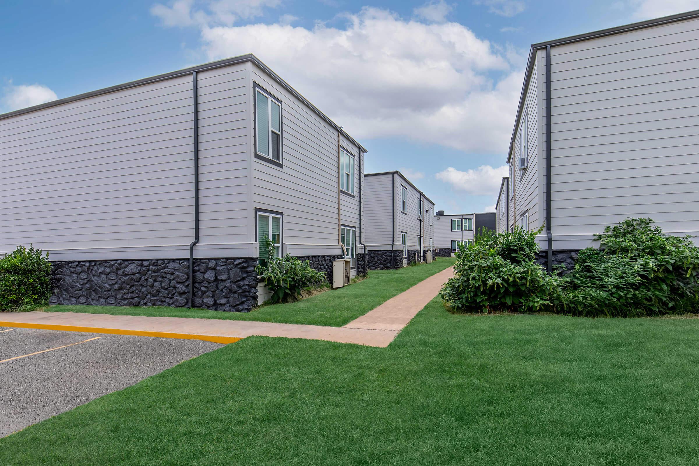 View of a modern residential area featuring gray modular buildings on either side of a paved walkway. Lush green grass and small plants line the pathway, creating a pleasant and inviting atmosphere. The sky is partly cloudy, contributing to a bright and open feel.